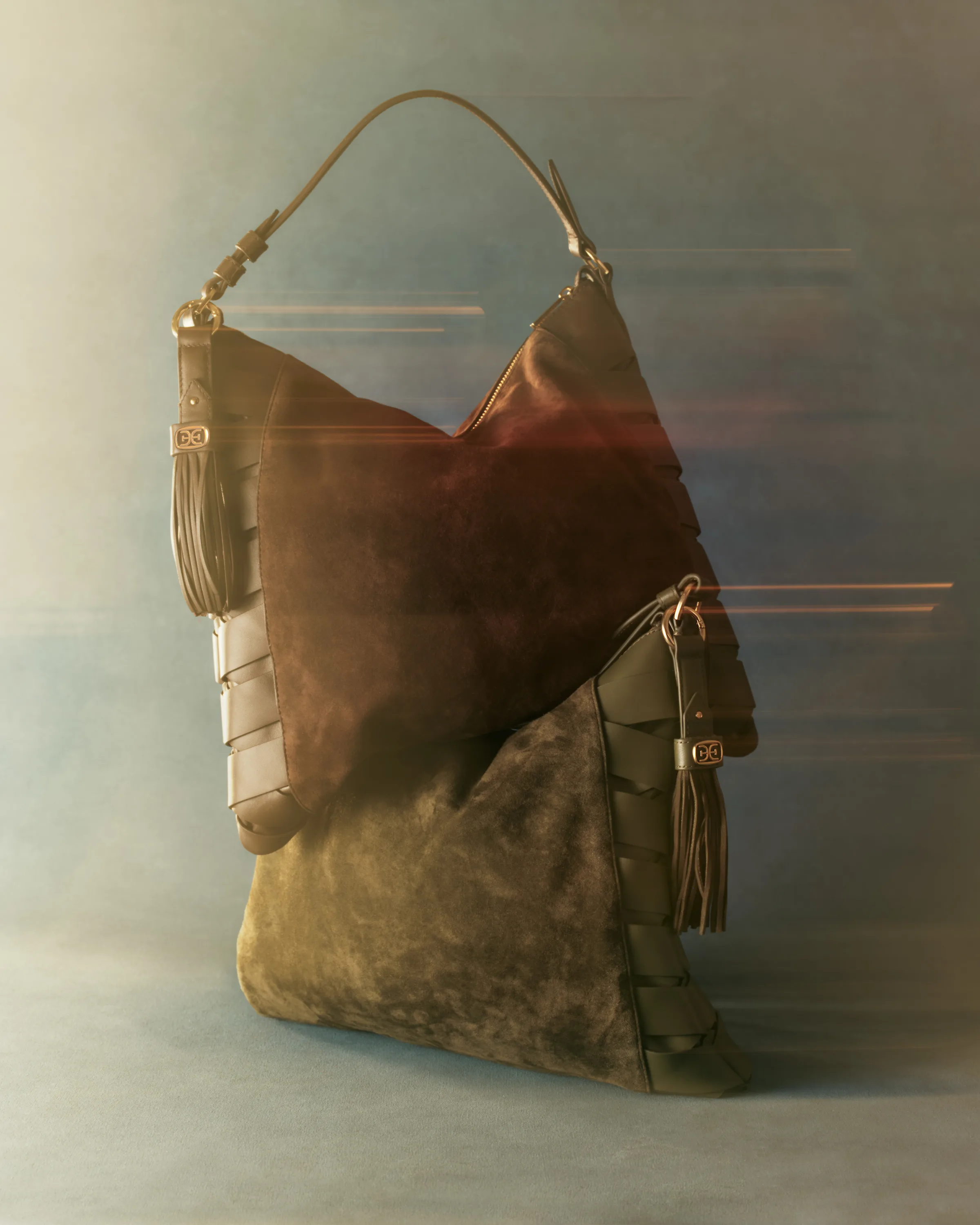 Two suede hobo bags in brown and olive with woven leather sides and tassels, stacked against a blurred blue backdrop, for Sam Edelman.