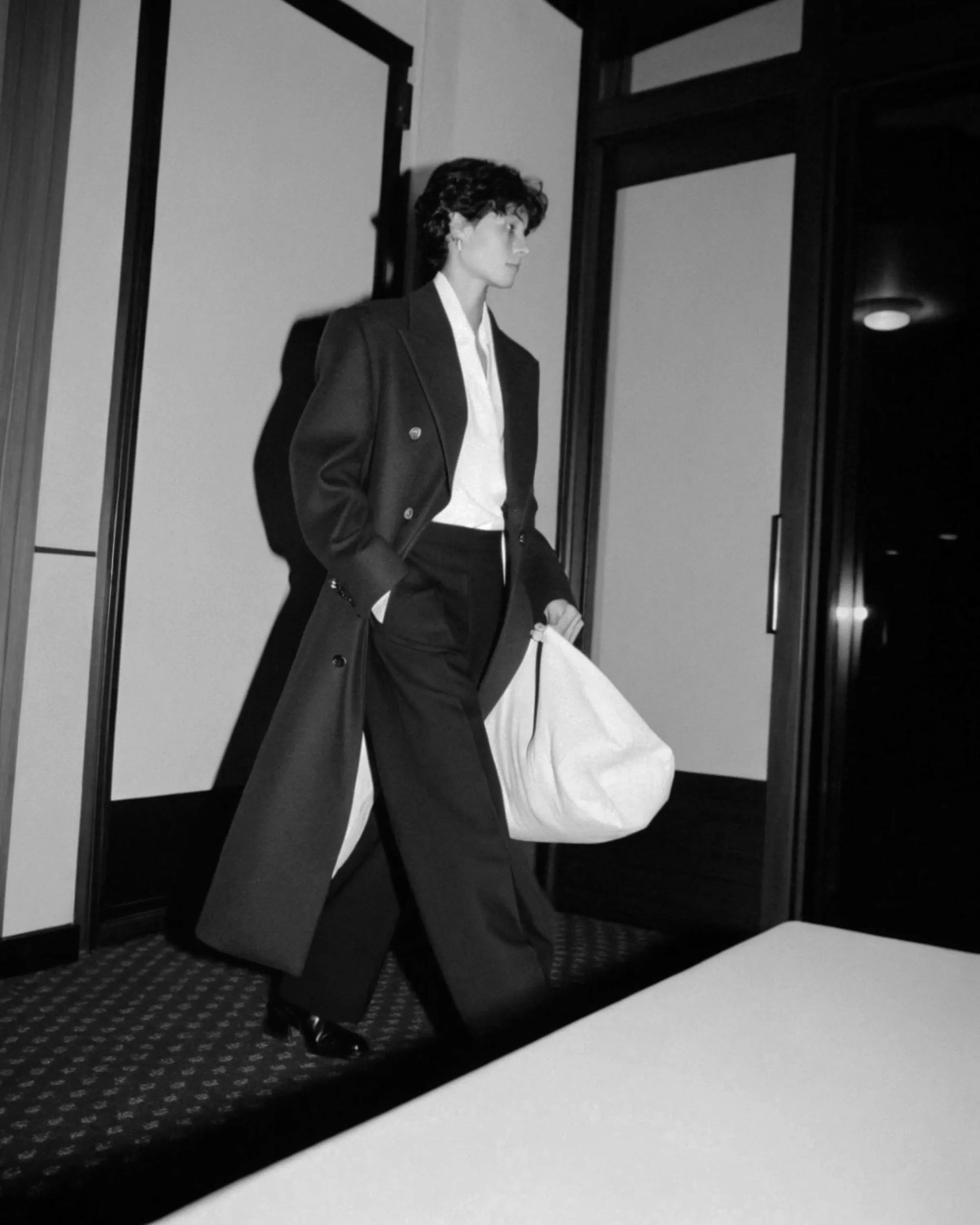 Black-and-white AI image of a model in a long dark coat carrying a white tote down a hotel corridor. Fursac AI Concept.