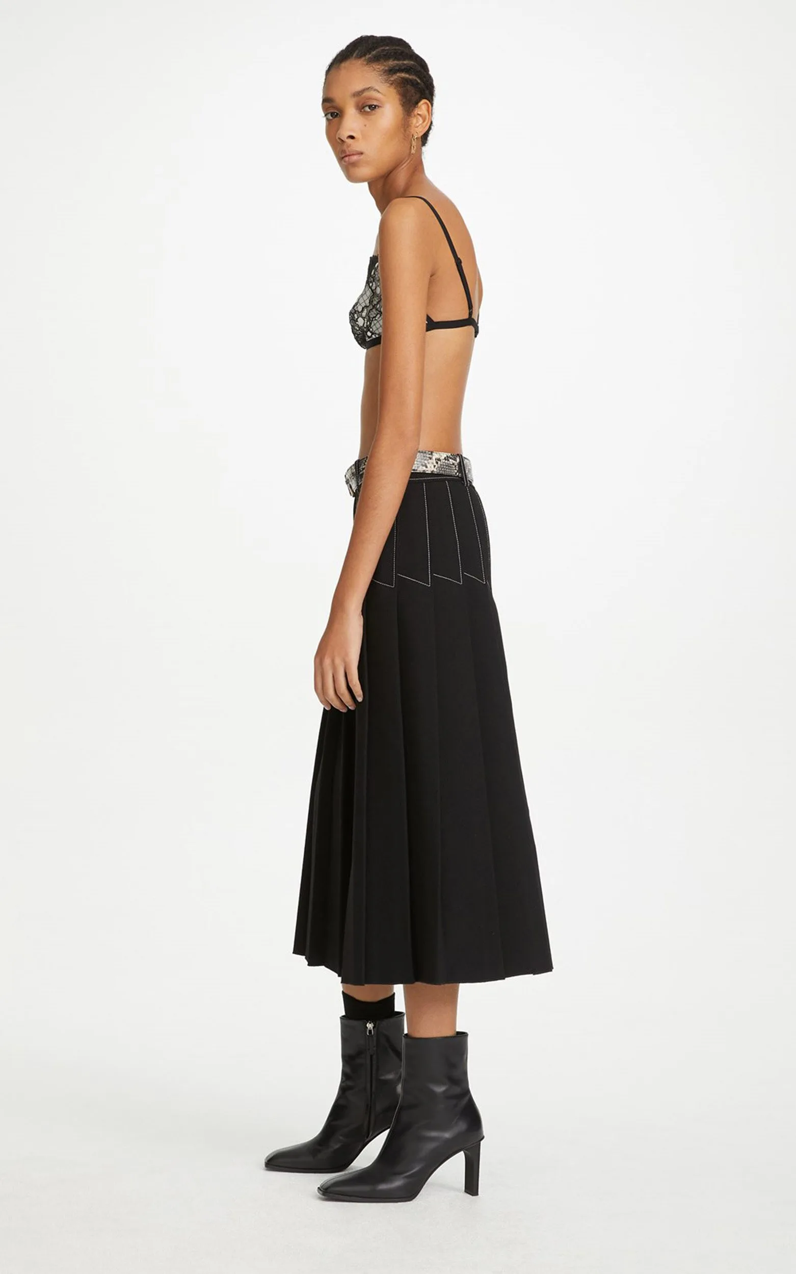 Model in profile wearing a black pleated midi skirt with contrast stitching, printed bra top and black boots, for Dion Lee.