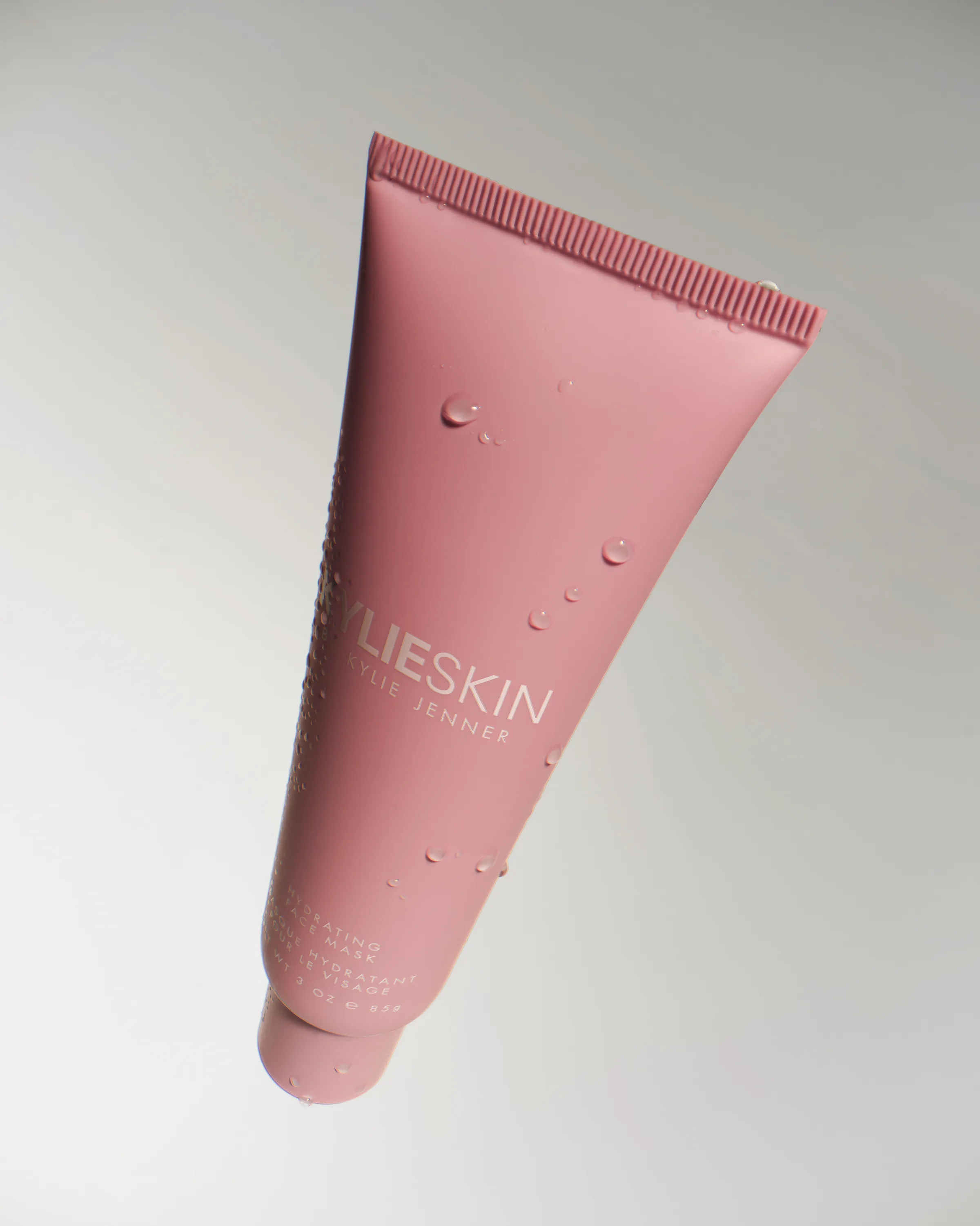 Pink Kylie Skin hydrating face mask tube with water droplets, floating against a soft grey background, for Kylie Cosmetics.