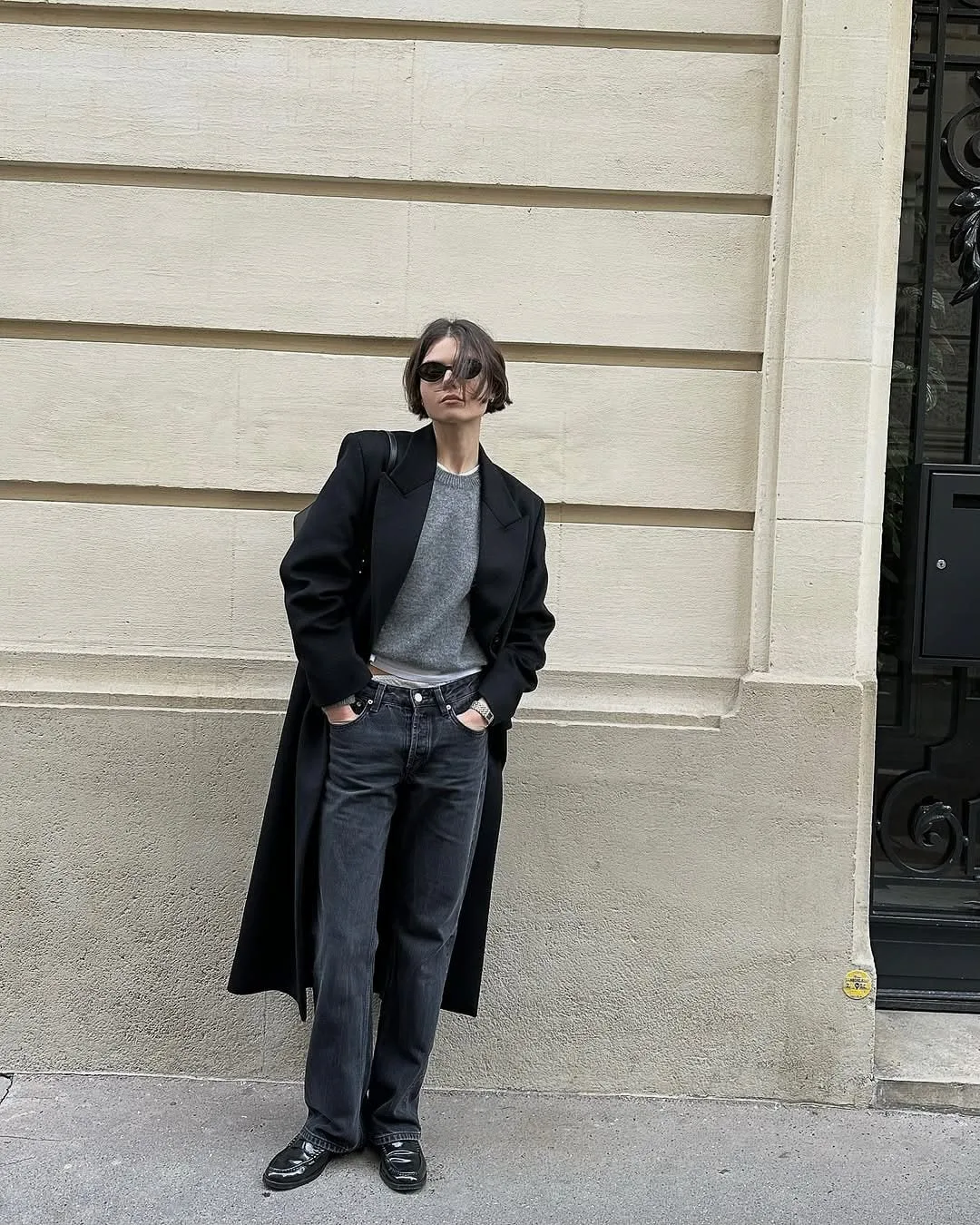 Woman in a long black coat, grey sweater, dark jeans and loafers posing against a cream stone wall on a city street.