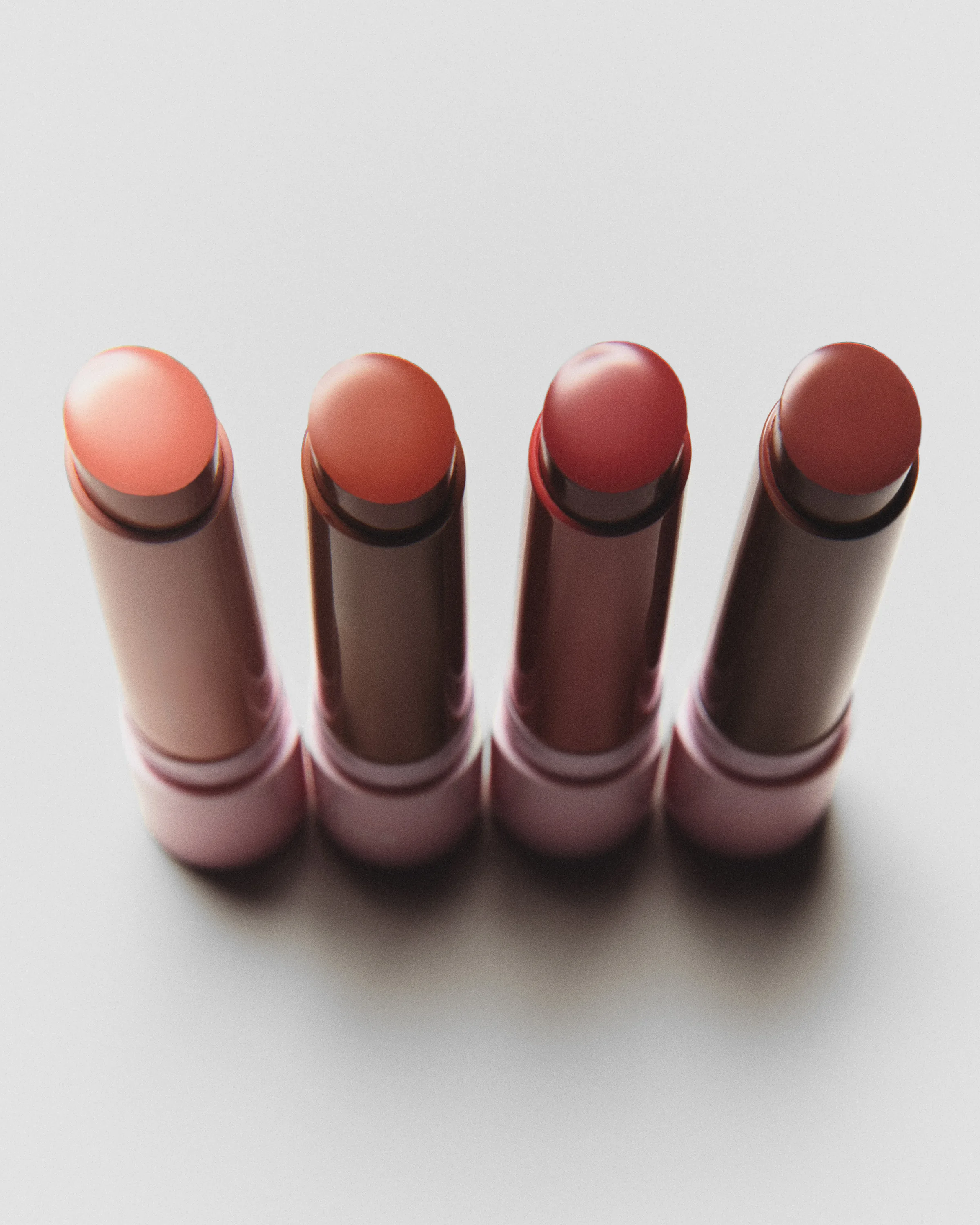 Four uncapped lip balms in peach, coral, rose and mauve shades standing in a row on a cream background, for Kylie Cosmetics.