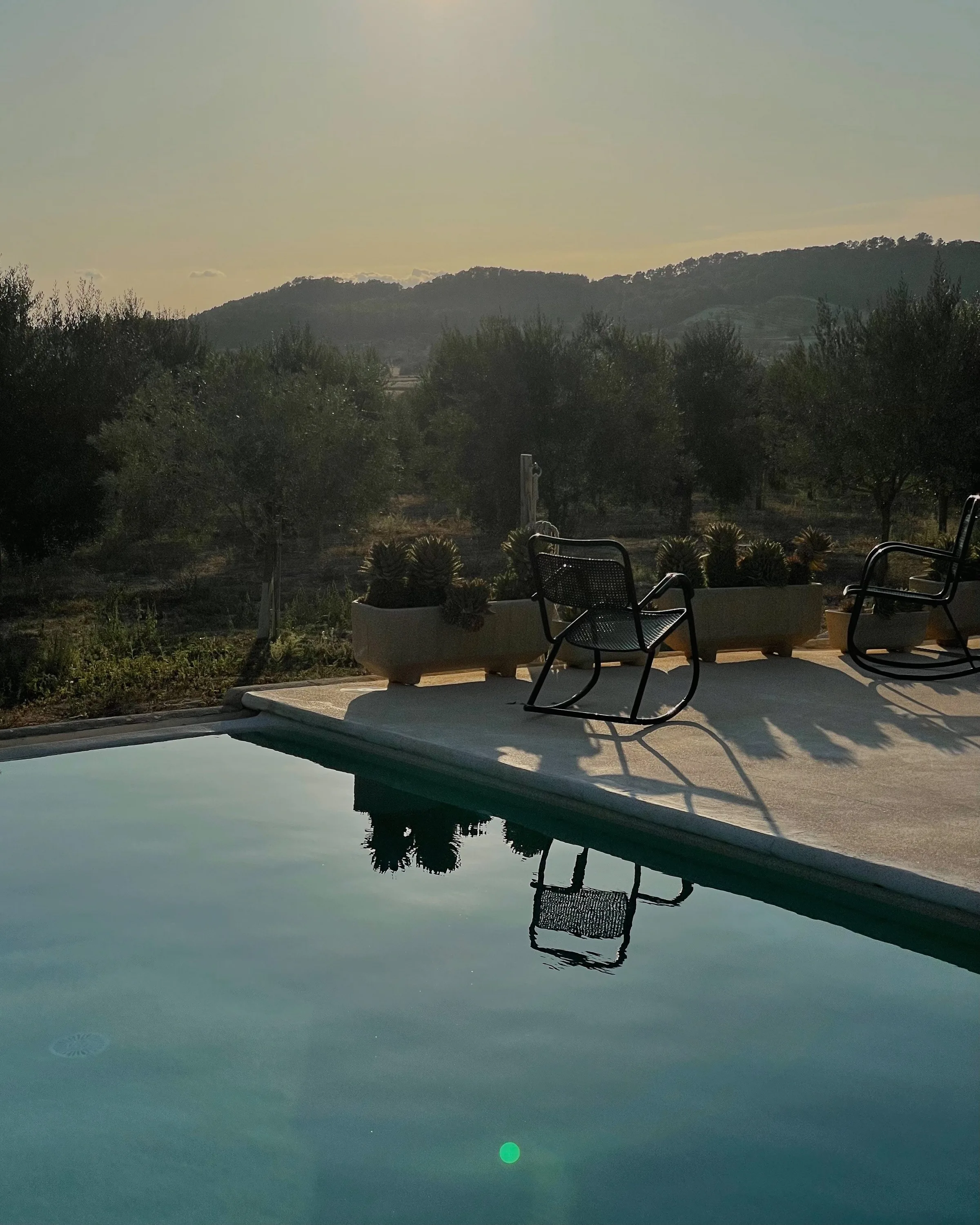 Black metal rocking chairs beside a still pool at dusk, with cacti planters, olive trees and hills behind.