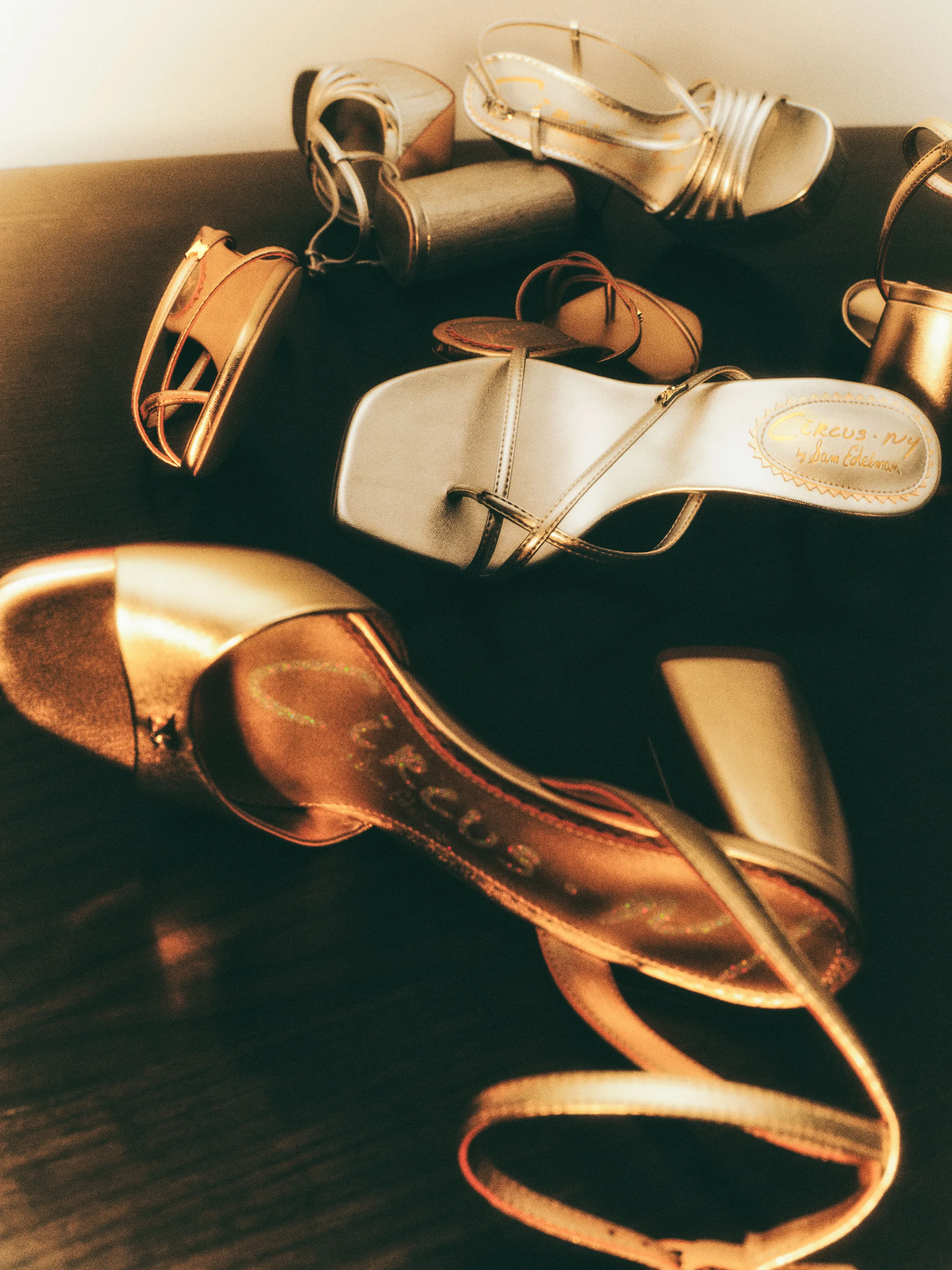 Gold and silver metallic strappy sandals scattered on a dark wooden surface in warm light, for Circus NY.