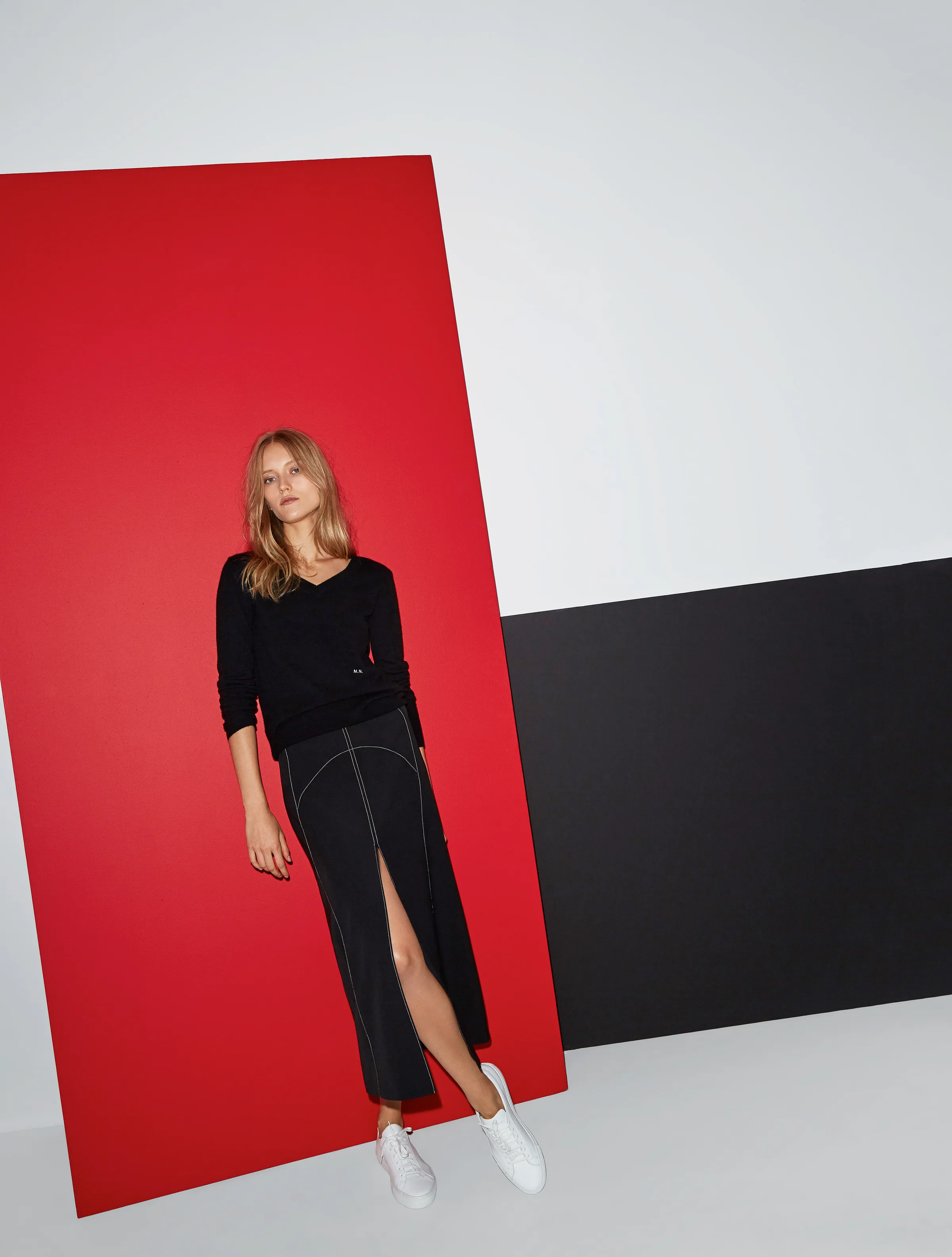 Model in a black V-neck sweater and contrast-stitched slit skirt leaning on a red panel, for Misha Nonoo.
