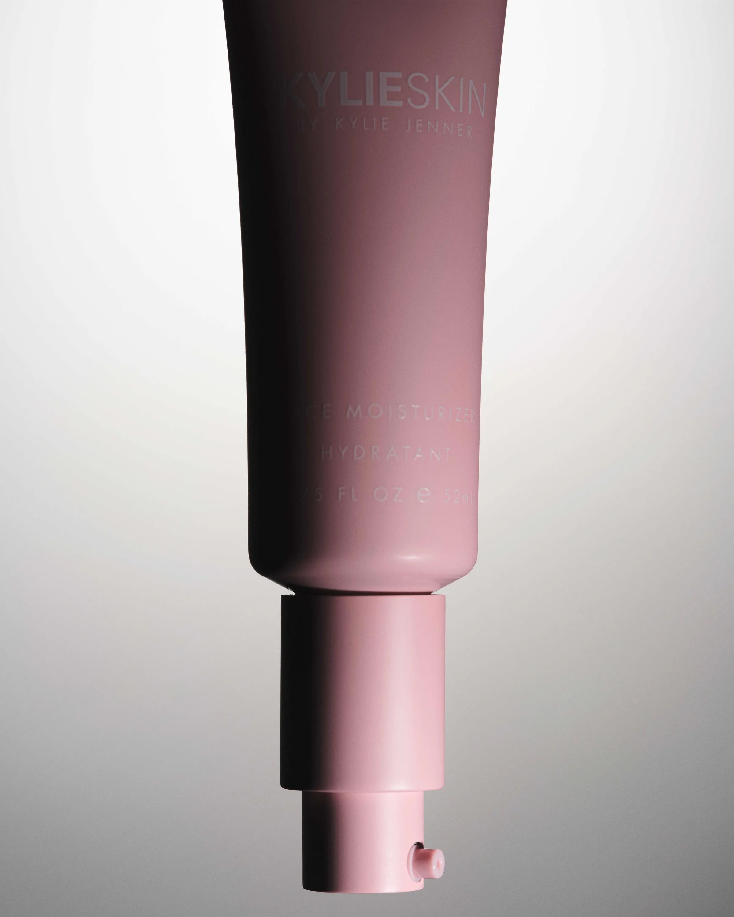 Still life of a pink Kylie Skin moisturizer tube with pump, hanging upside down against a grey gradient, for Kylie Cosmetics.