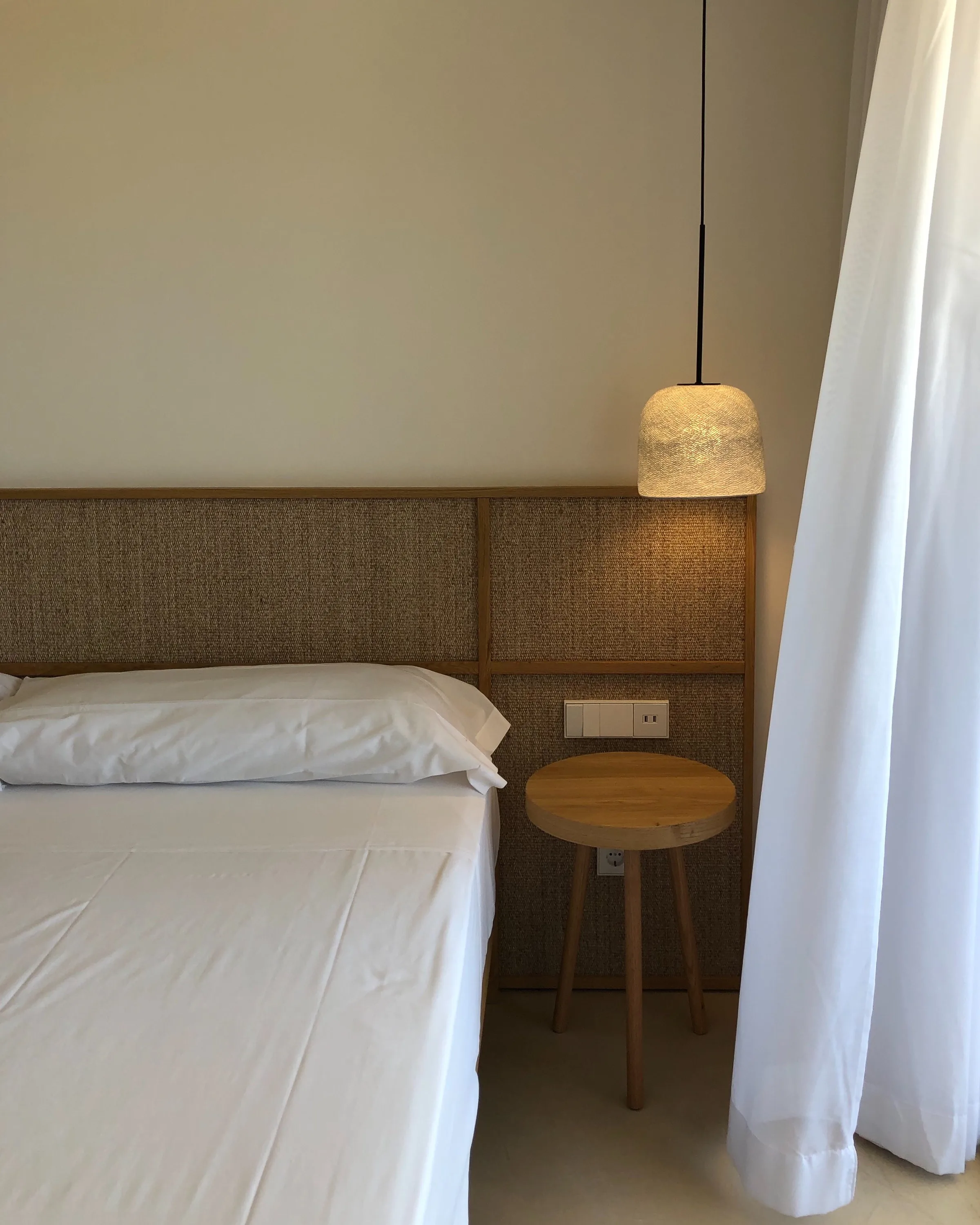 Bedroom corner with white bedding, woven rattan headboard, round wooden side stool and pendant lamp beside sheer curtains.