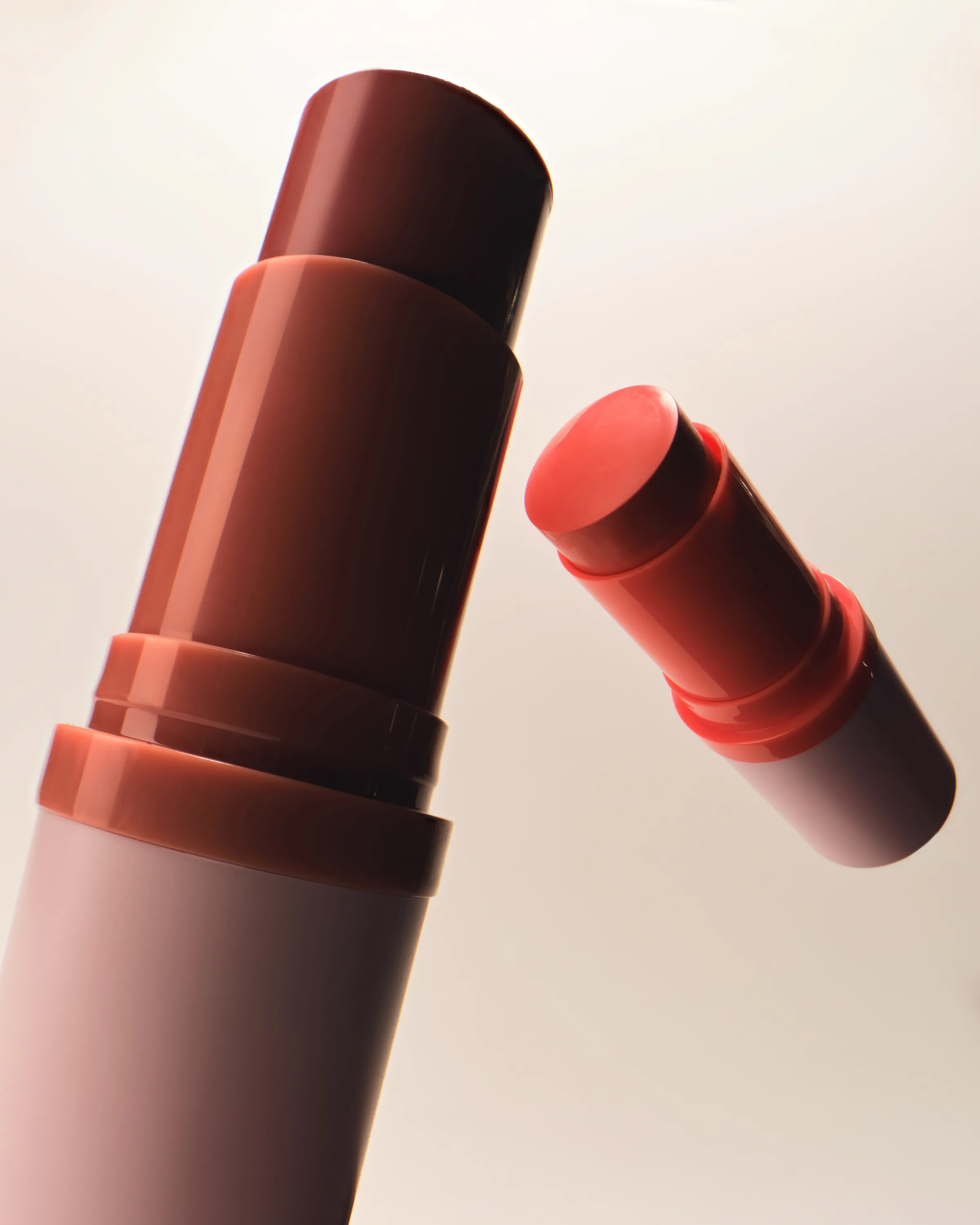 Two blush sticks in terracotta and coral, uncapped and angled against a soft cream background, for Kylie Cosmetics.