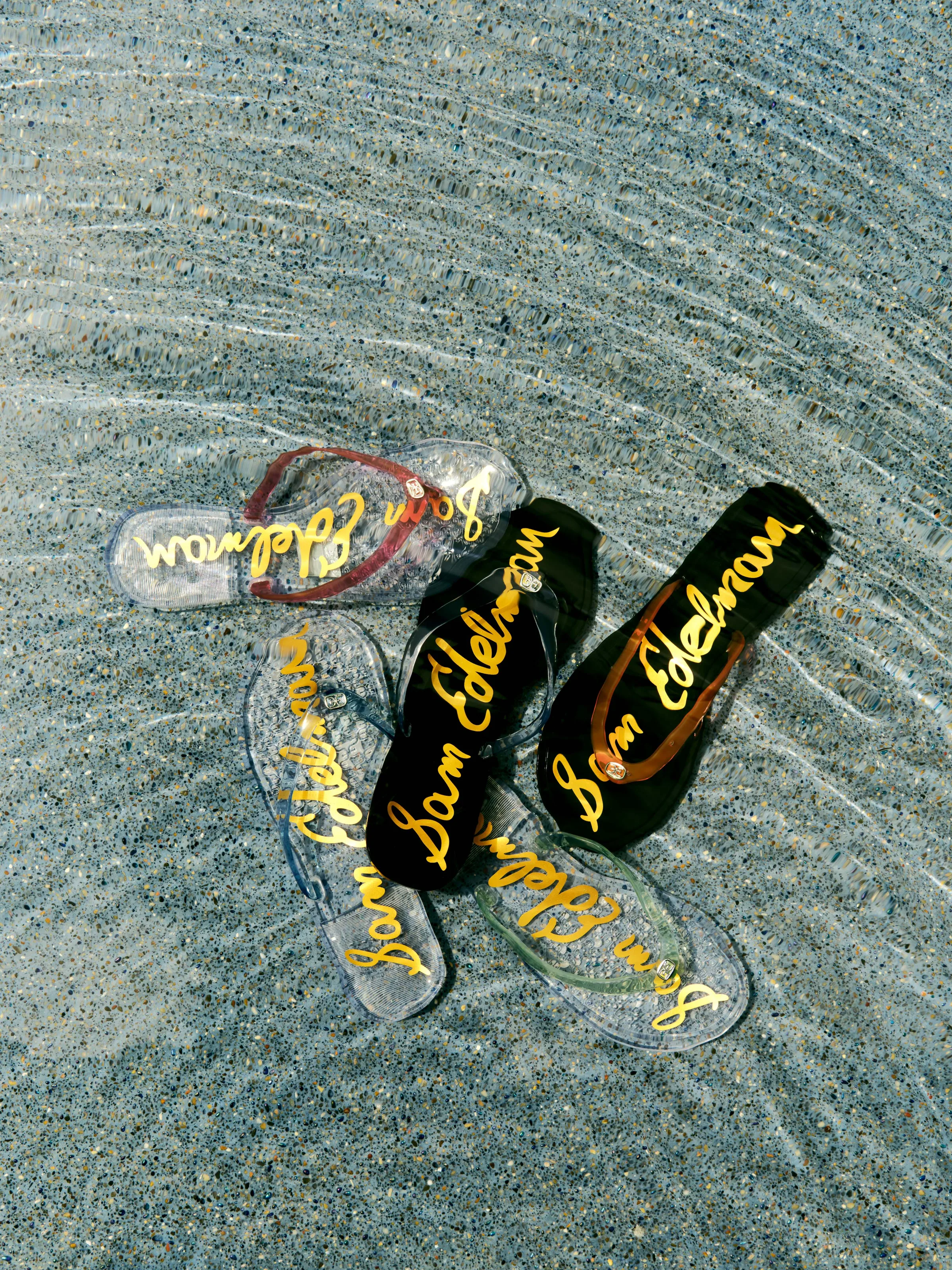Clear, black and tinted jelly flip-flops with gold script logos lying in shallow rippling water, for Sam Edelman.