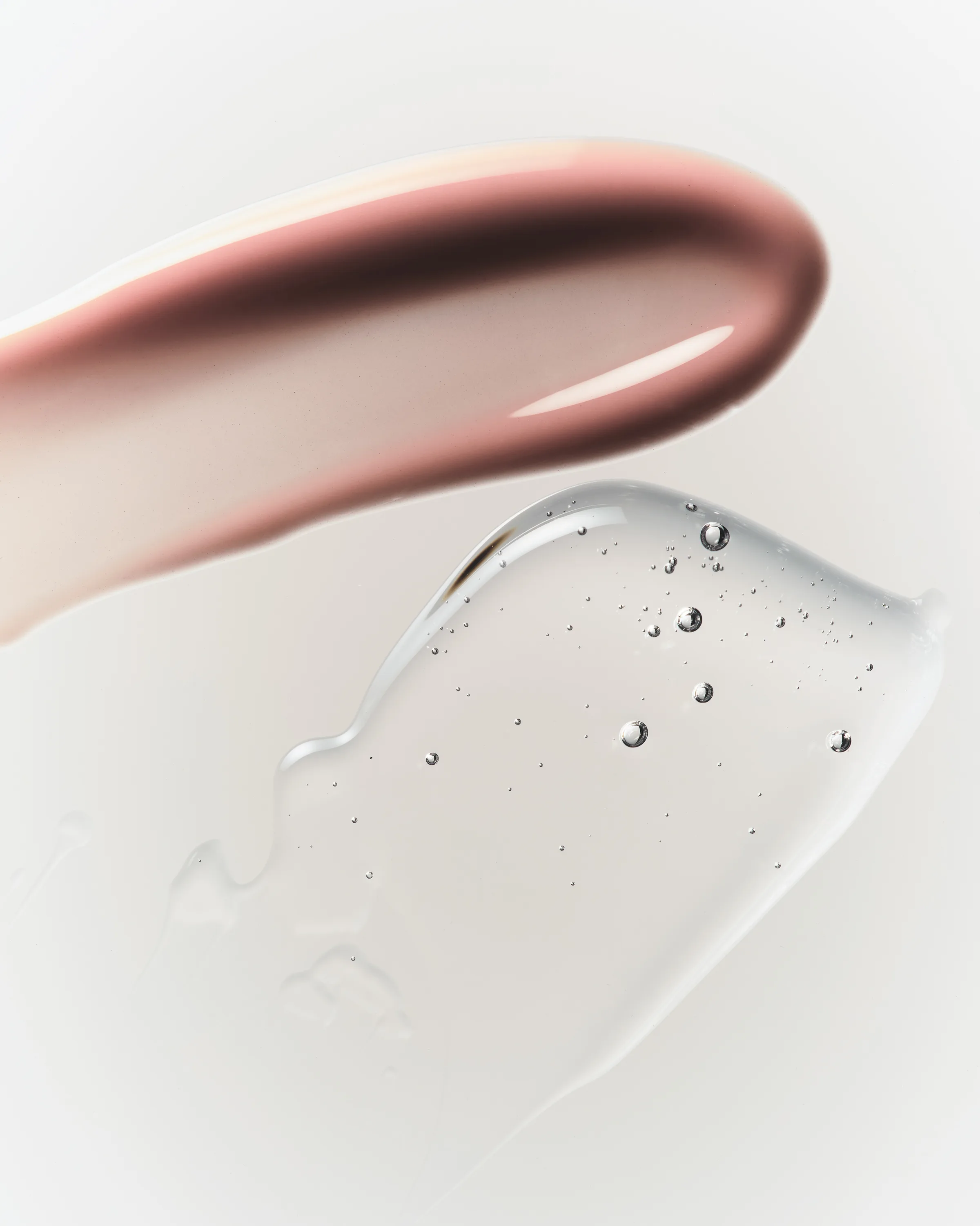 Swatches of rosy pink and clear lip oil with tiny bubbles on a pale grey surface, for Kylie Cosmetics.