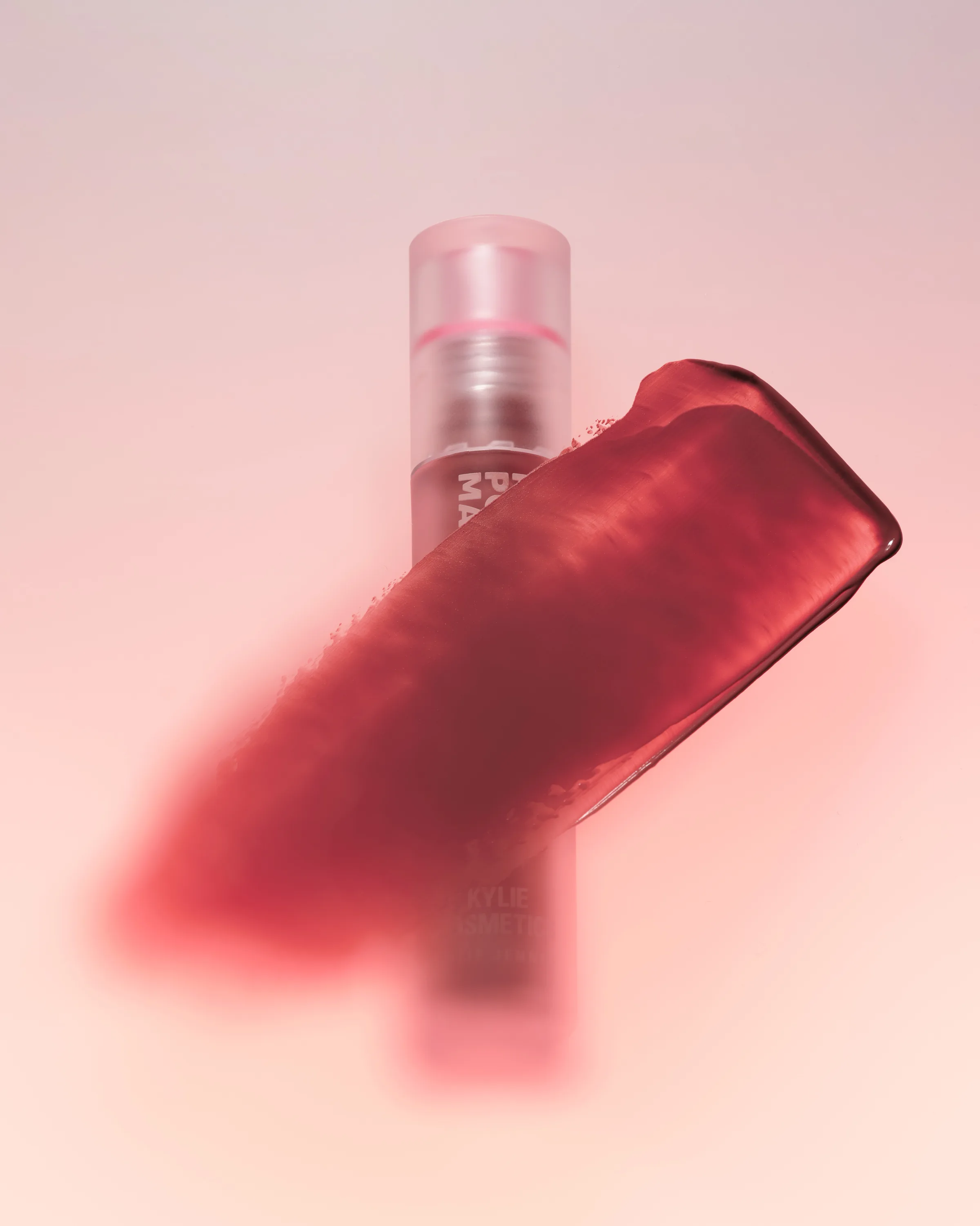Red lipstick swatch smeared across a blurred Love Letter lipstick tube on a soft pink background, for Kylie Cosmetics.