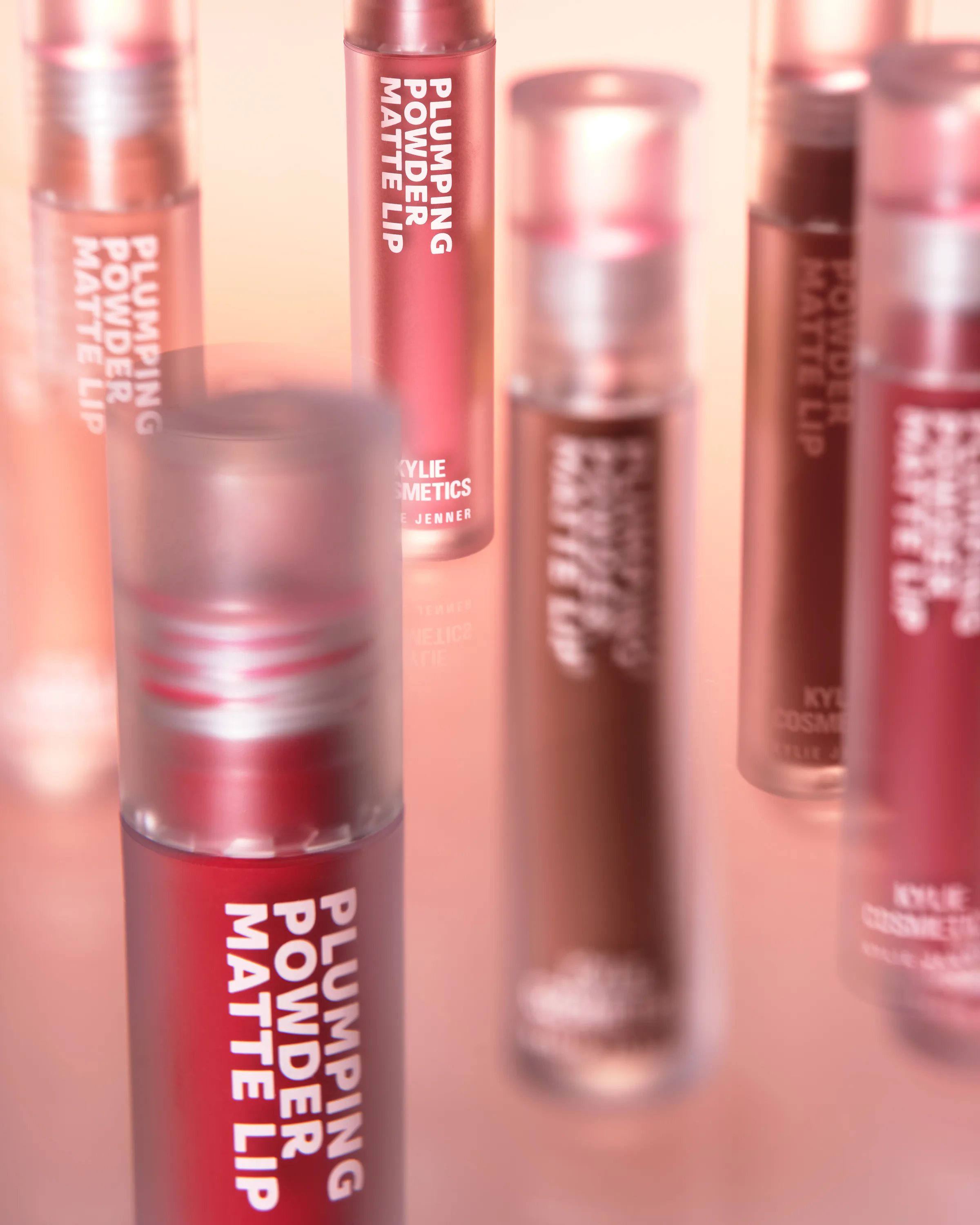 Plumping Powder Matte Lip tubes in red, rose and brown shades on a reflective peach surface, for Kylie Cosmetics.
