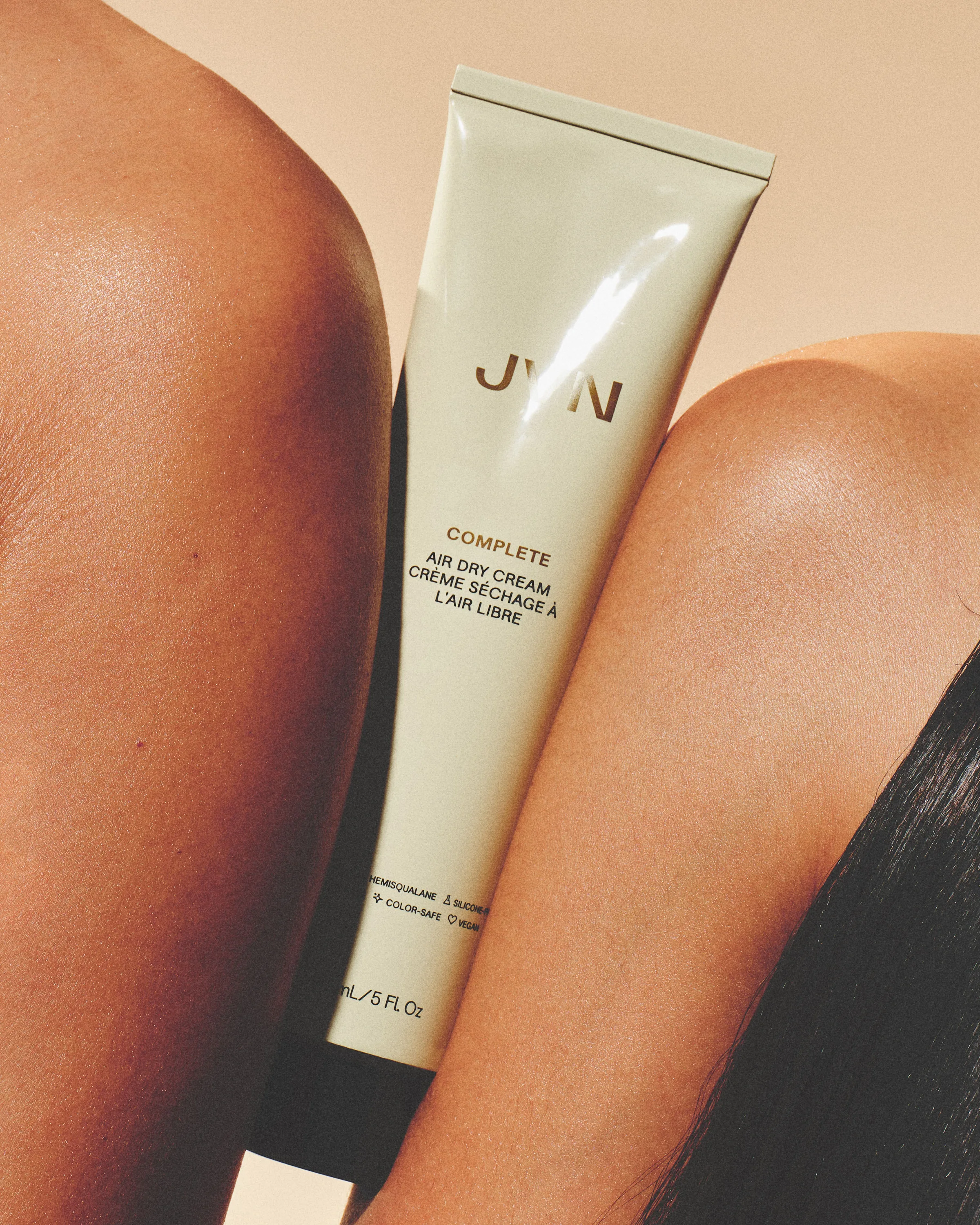 Cream tube of Complete Air Dry Cream held between two bare shoulders against a warm beige backdrop, for JVN.