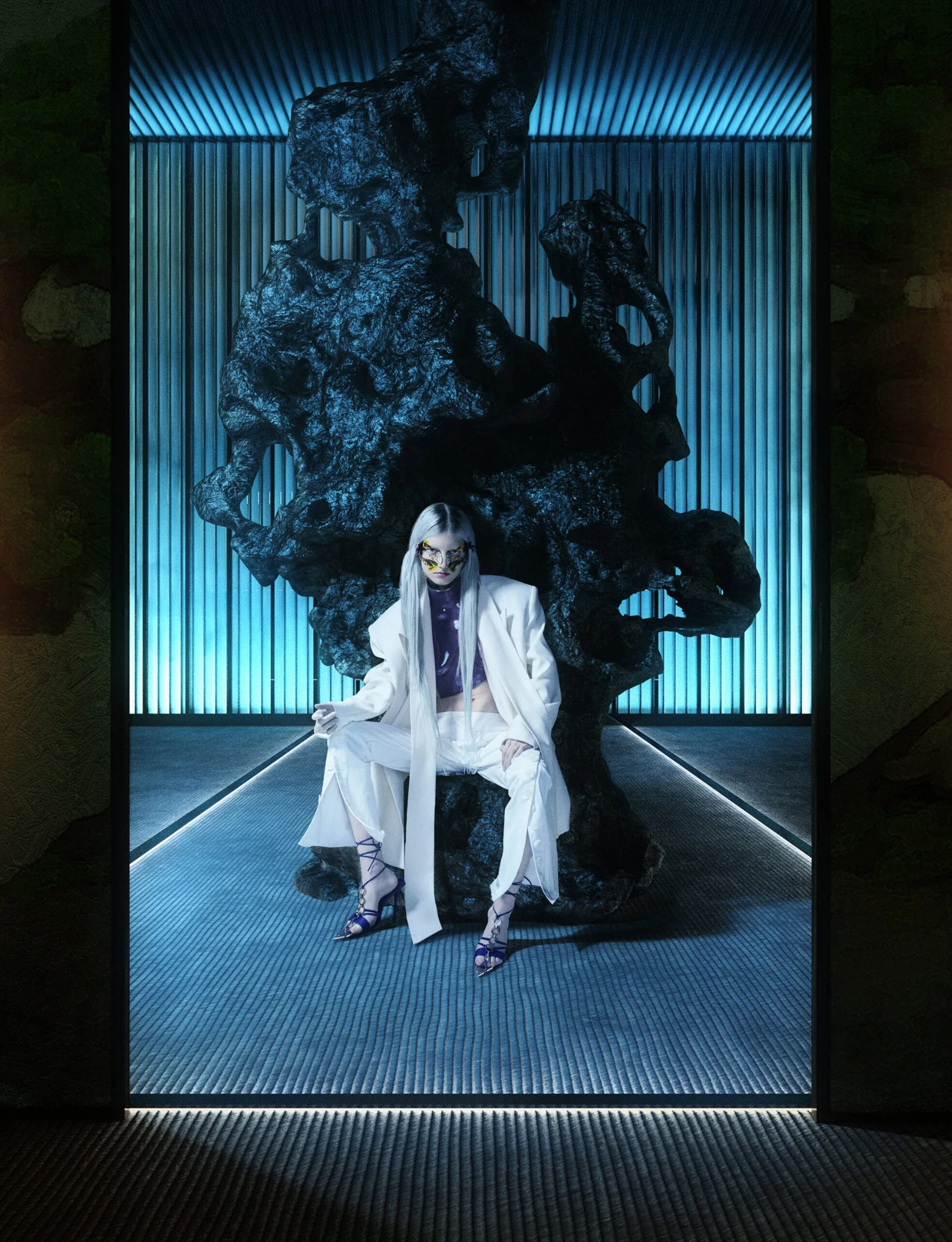 Model with silver hair and gold mask in a white suit, seated before a dark rock sculpture in blue-lit room, for Dazed & Confused.