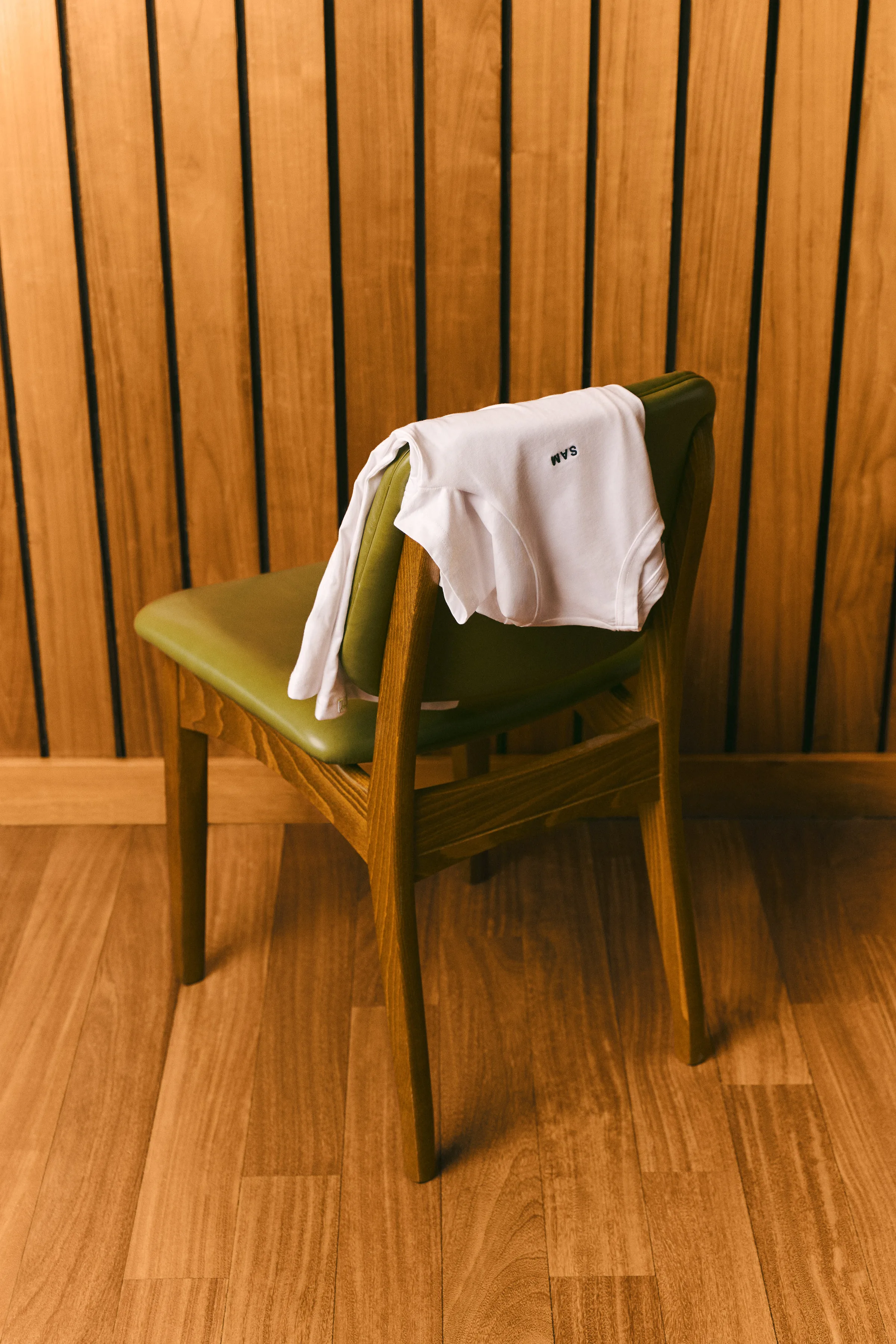 White t-shirt draped over an olive leather wooden chair against slatted wood paneling, for Sam Edelman.
