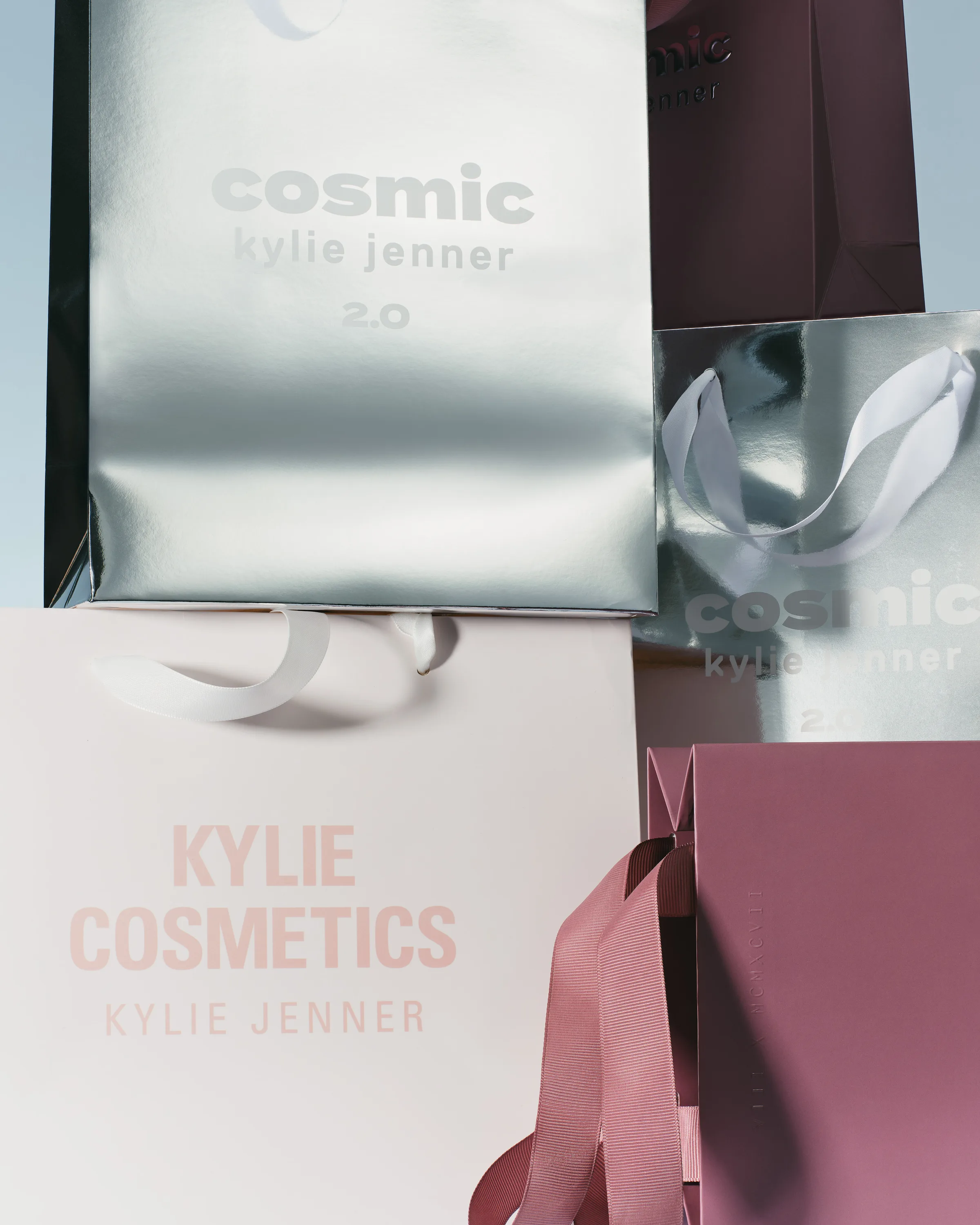 Stacked silver Cosmic 2.0, pale pink and mauve shopping bags with ribbon handles against a pale blue sky, for Kylie Cosmetics.