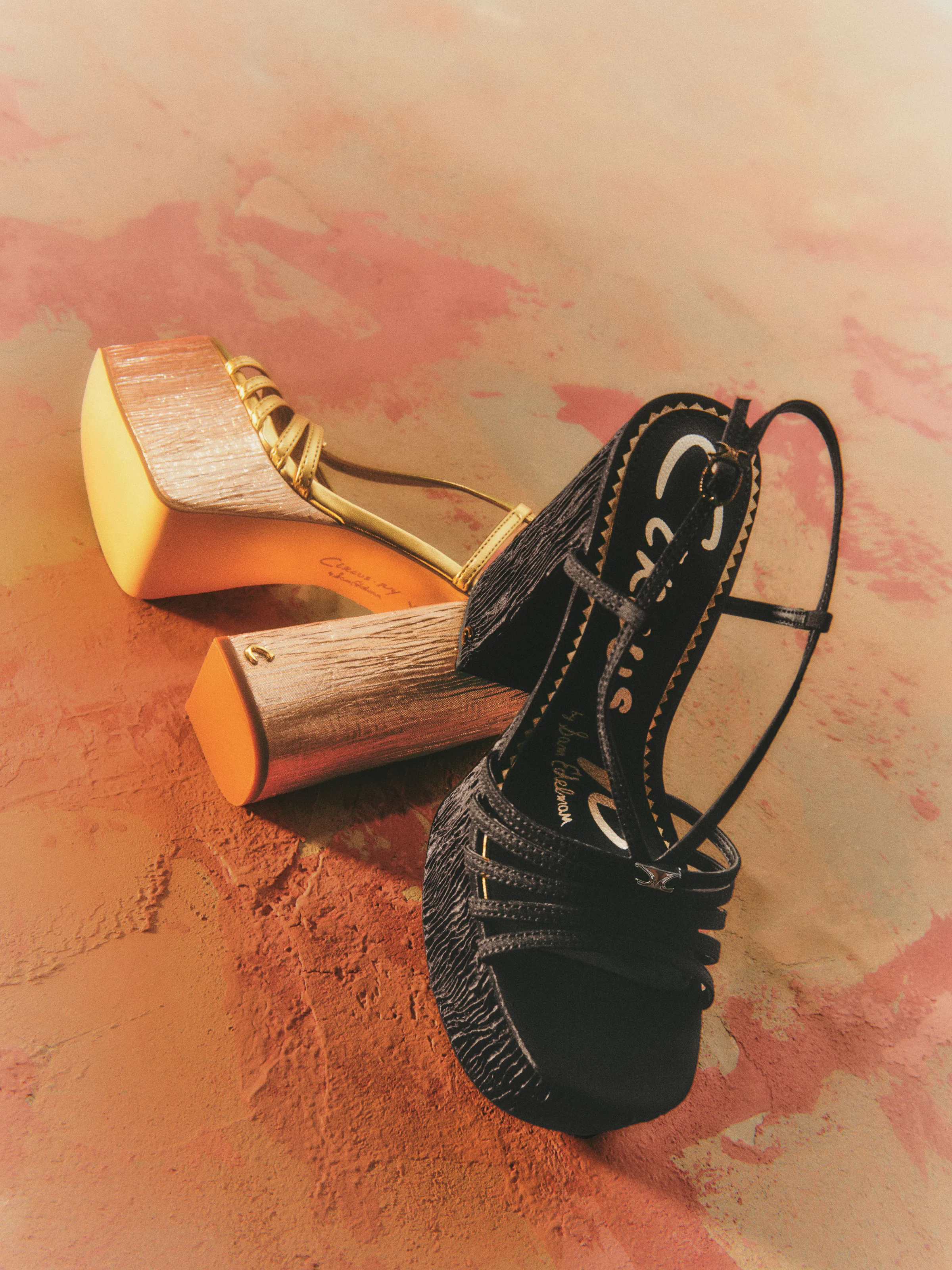 Gold and black strappy platform sandals with textured wooden heels on a dusty terracotta surface, for Circus NY.