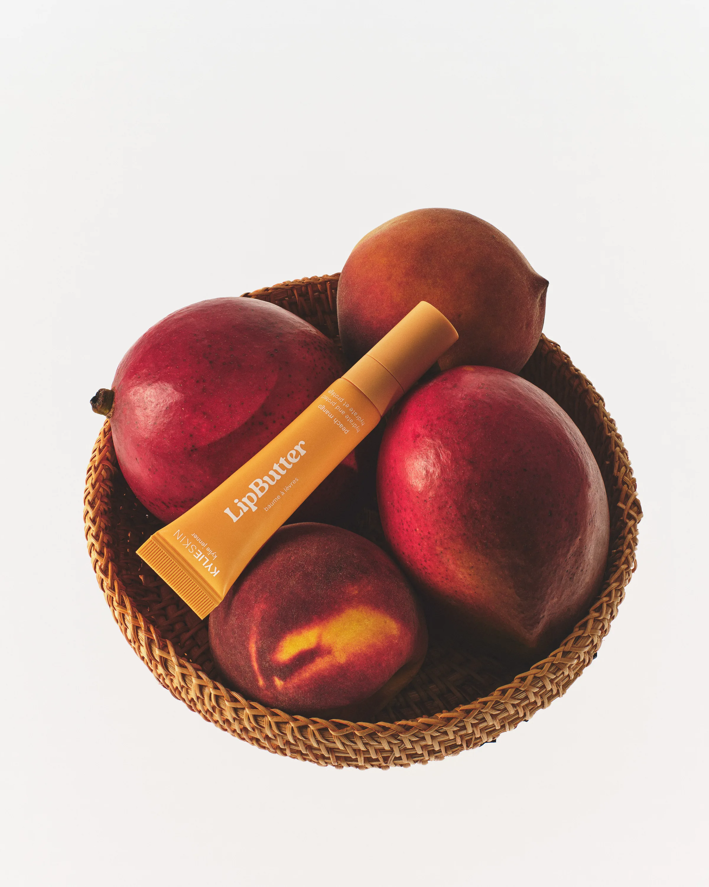 Orange Lip Butter tube resting on mangoes and a peach in a woven basket on white, still life, for Kylie Cosmetics.