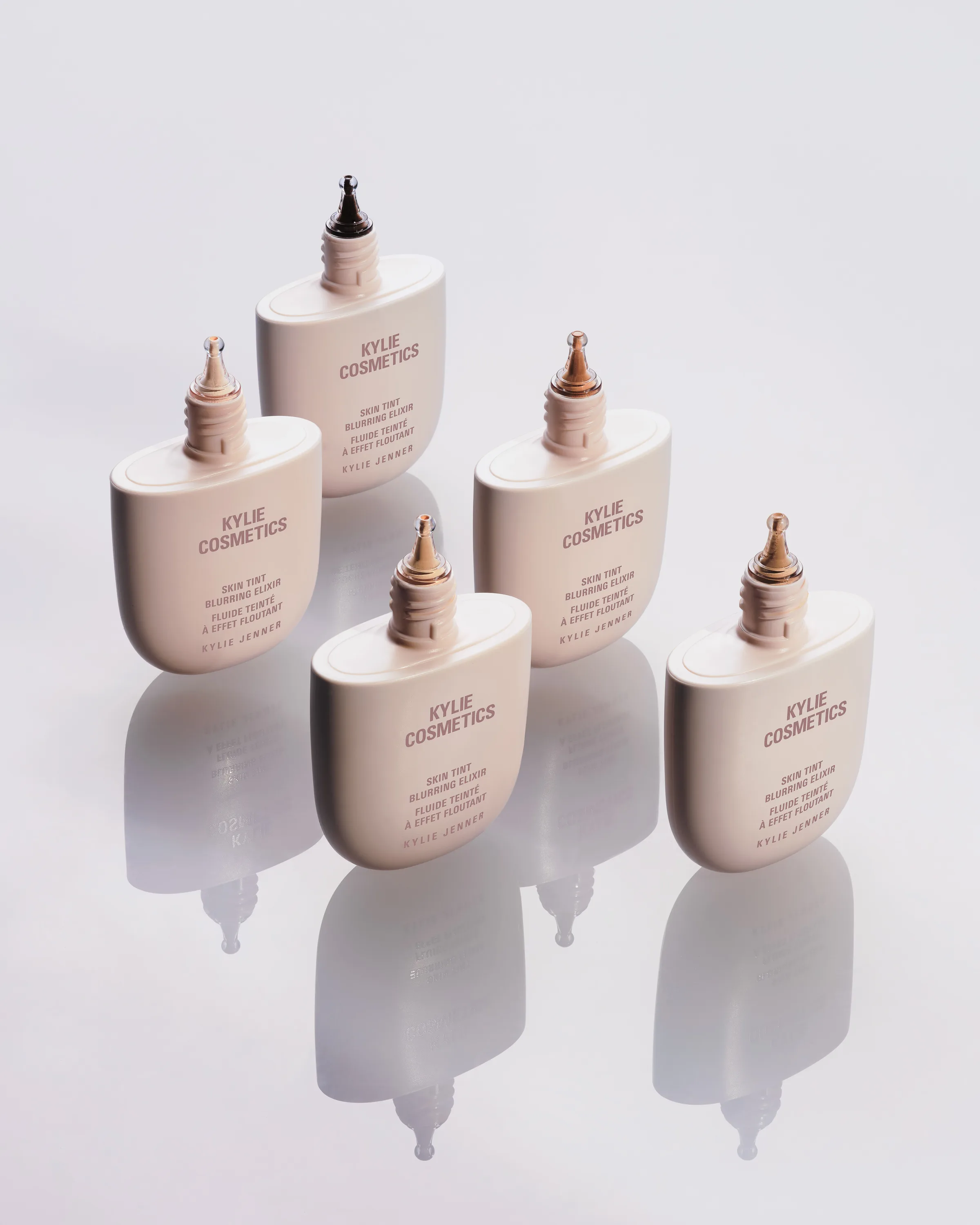 Five Skin Tint Blurring Elixir bottles in different shades standing on a reflective white surface, for Kylie Cosmetics.