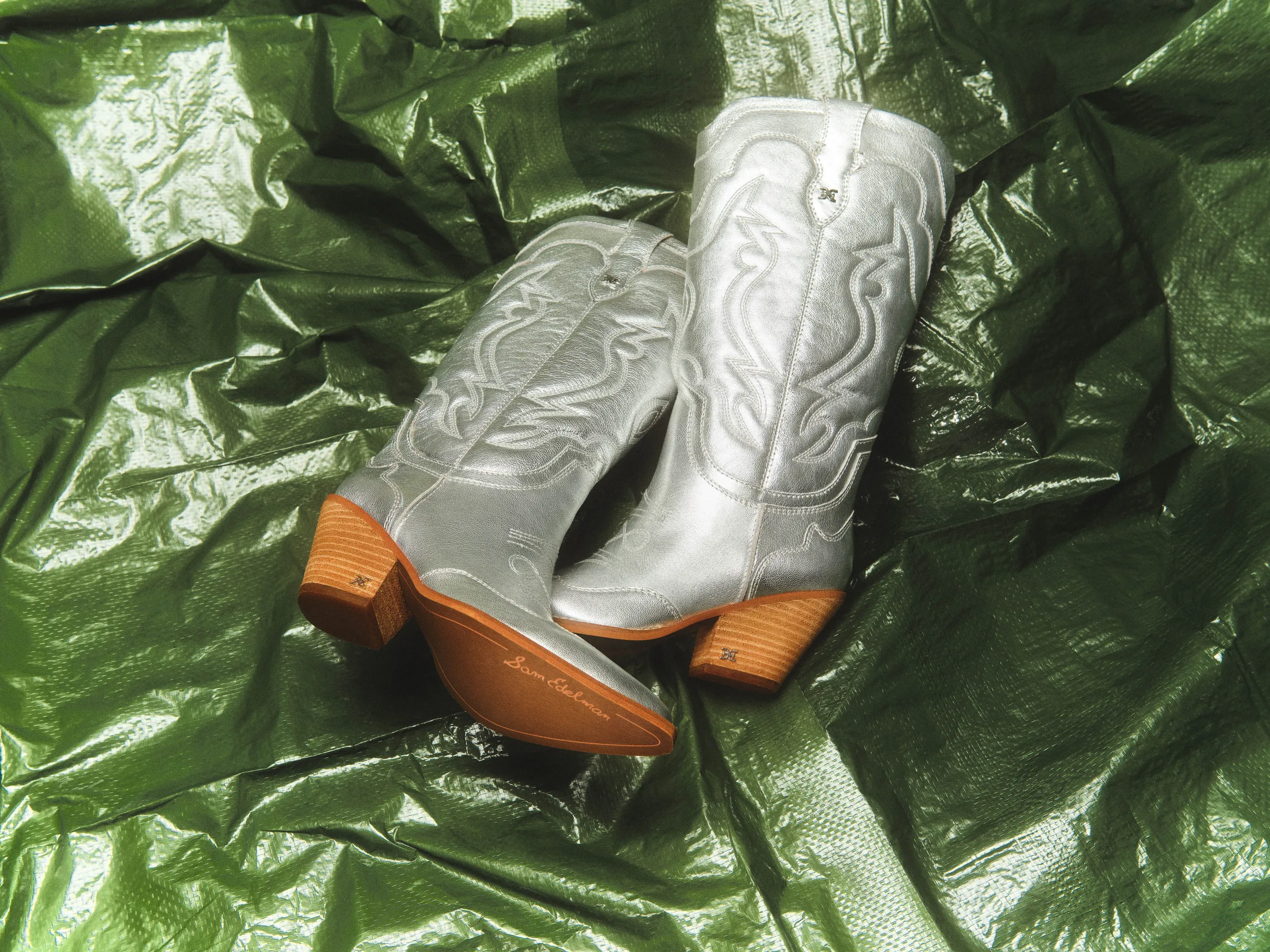 Metallic silver western boots with stitched shafts and stacked wood heels on crinkled green plastic, for Sam Edelman.