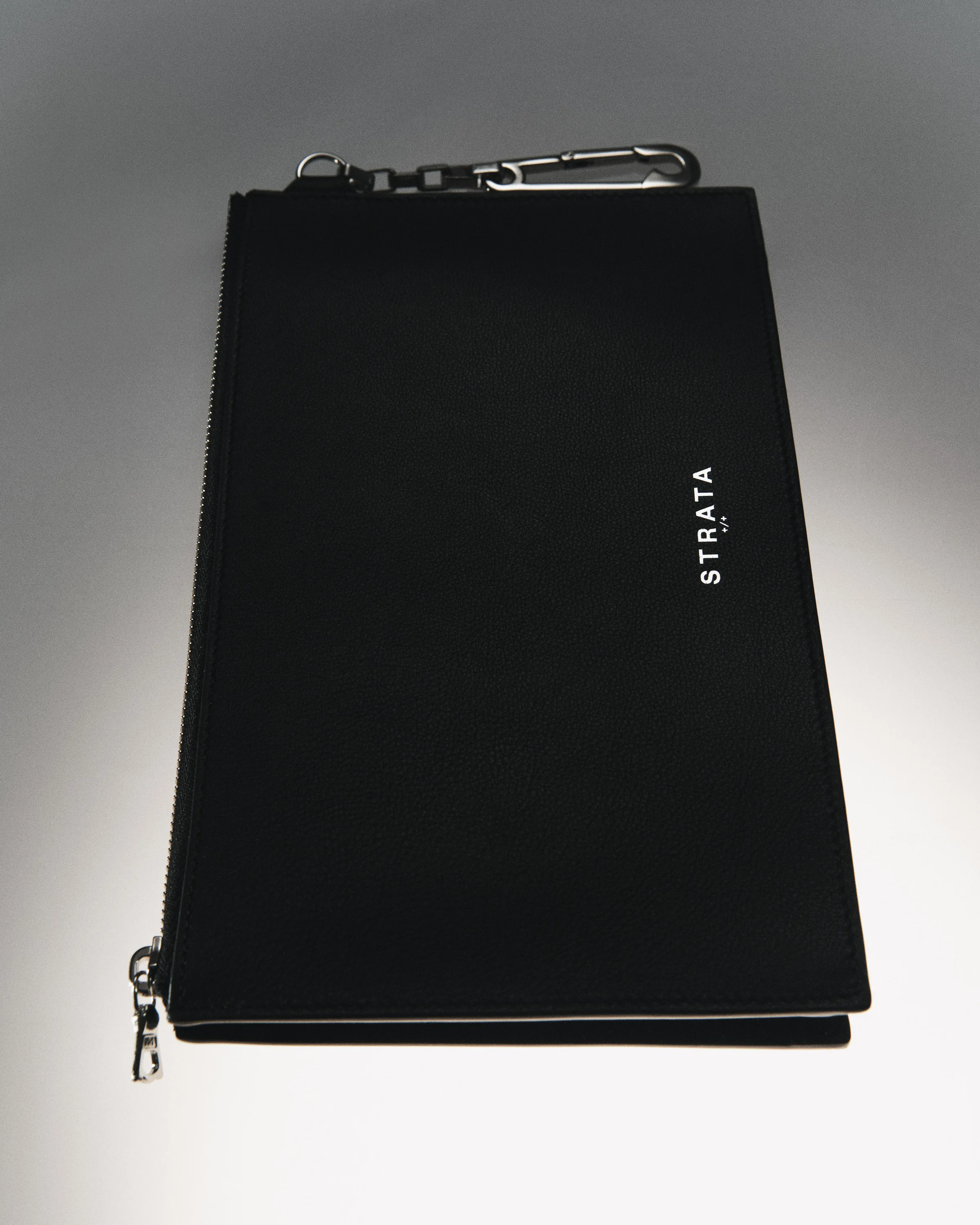 Black pebbled leather zip clutch with silver carabiner clip and white logo on a grey gradient backdrop, for Strata.