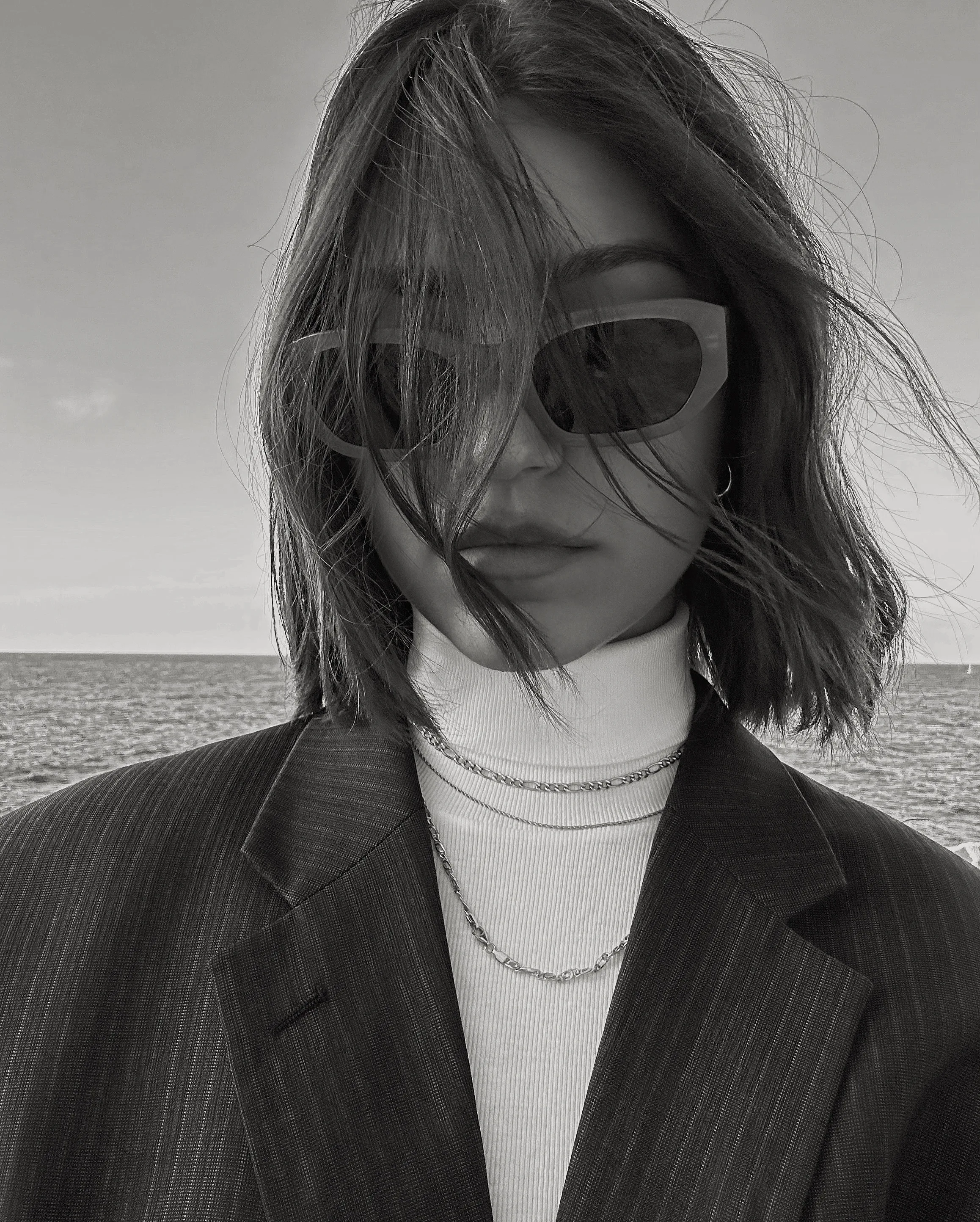Black and white selfie of a woman in sunglasses, turtleneck, chains and pinstripe blazer, hair blowing by the sea.