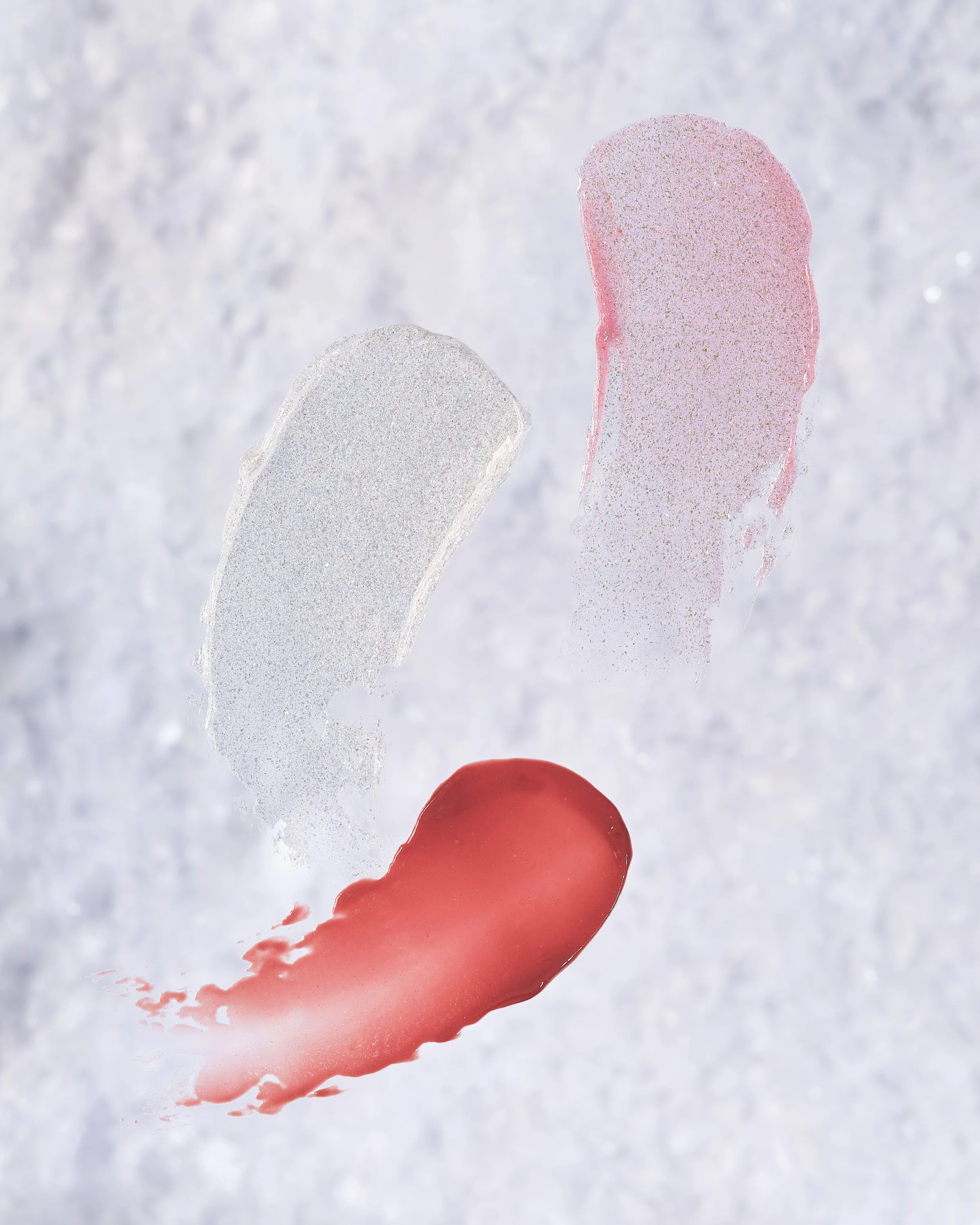 Three lip product swatches in clear glitter, pink shimmer and red on a snowy white surface, for Kylie Cosmetics.
