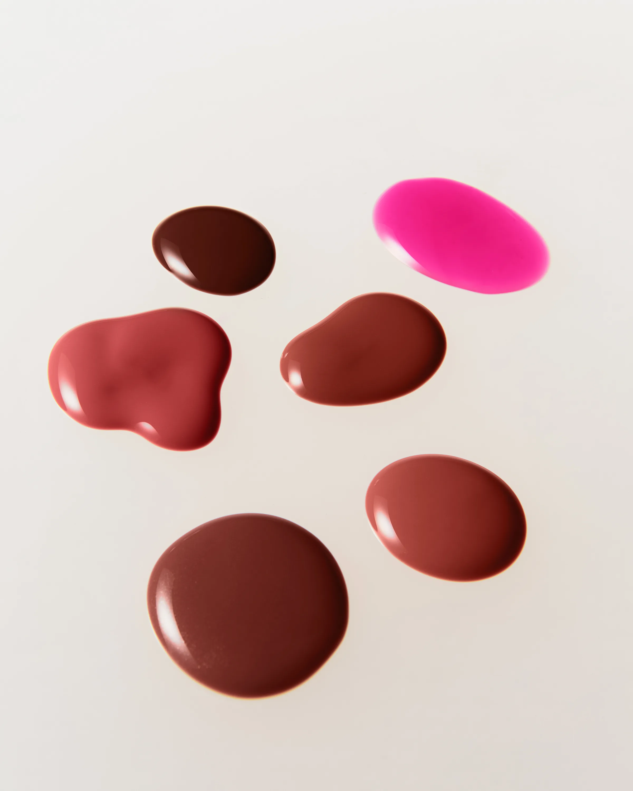 Six glossy lip glaze swatches in brown, rose and hot pink shades on a cream surface, for Kylie Cosmetics.