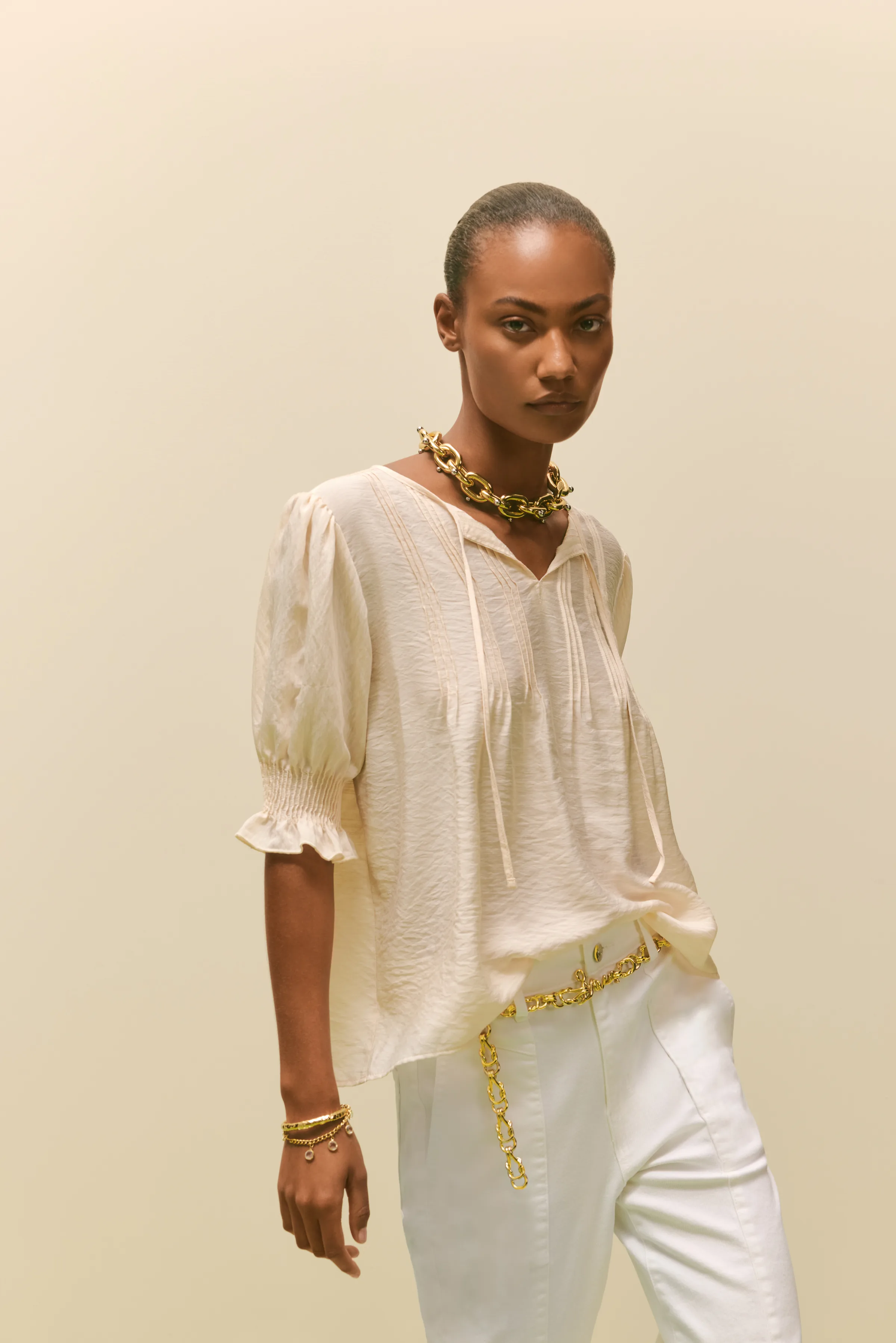 Model in a cream peasant blouse, white trousers, gold chain necklace and chain belt on a beige backdrop, for Sam Edelman.