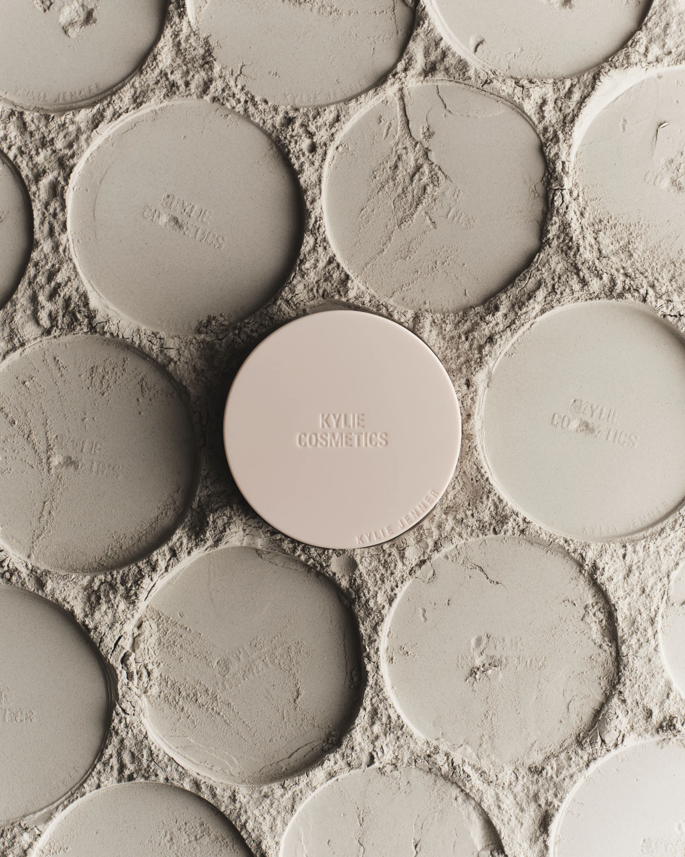 Closed beige powder compact centered among pressed powder discs on a textured powder surface, for Kylie Cosmetics.