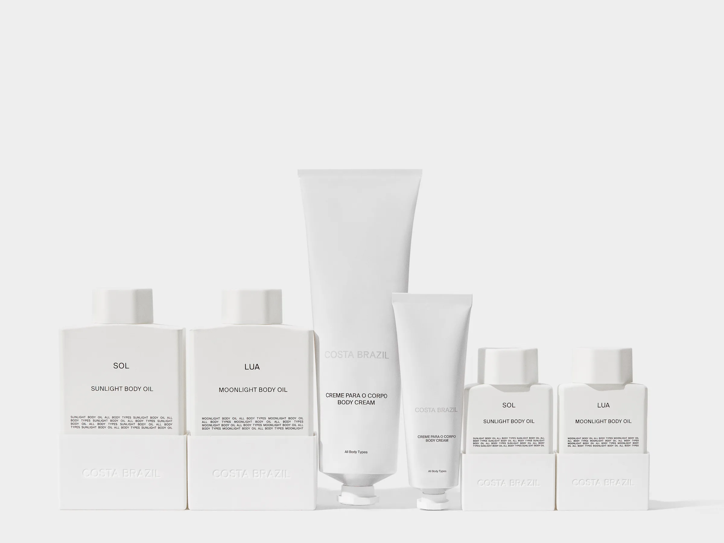 Lineup of white Sol and Lua body oil bottles and body cream tubes on a plain light grey background, for Costa Brazil.
