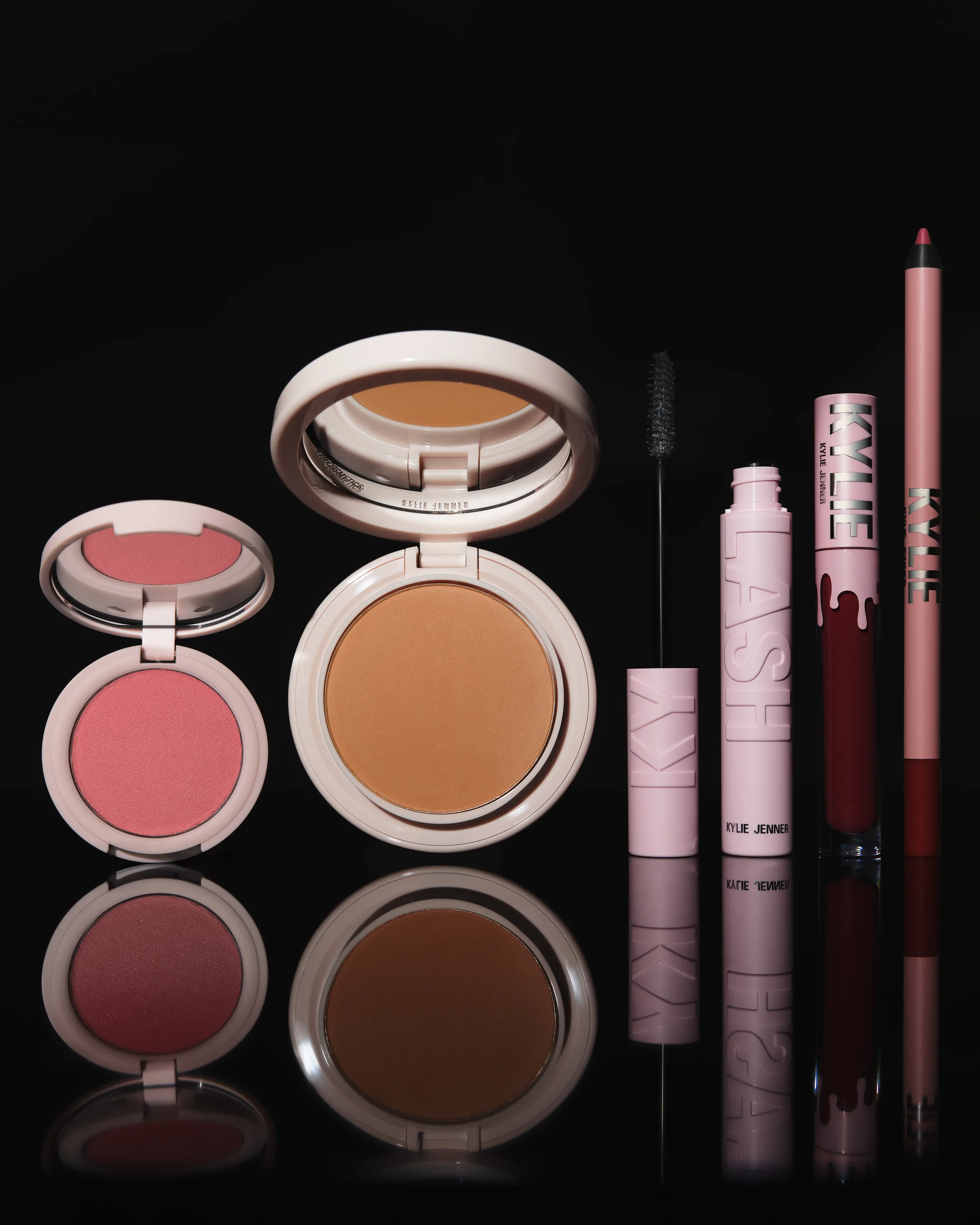 Pink compacts of blush and bronzer beside Kylash mascara, lip kit and liner reflected on glossy black, for Kylie Cosmetics.