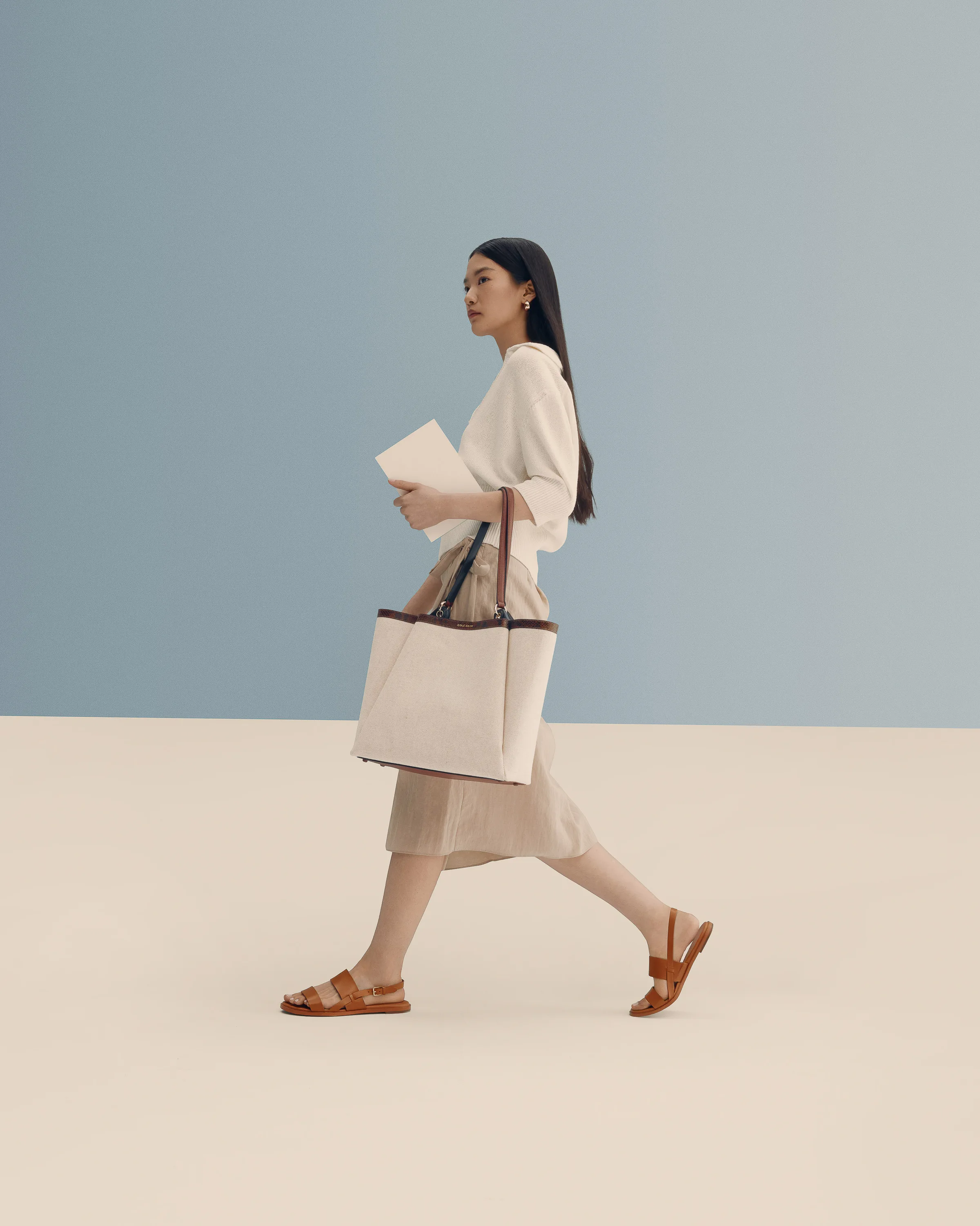 Model walking with a canvas tote and tan leather sandals against a blue and cream set, for Cole Haan.
