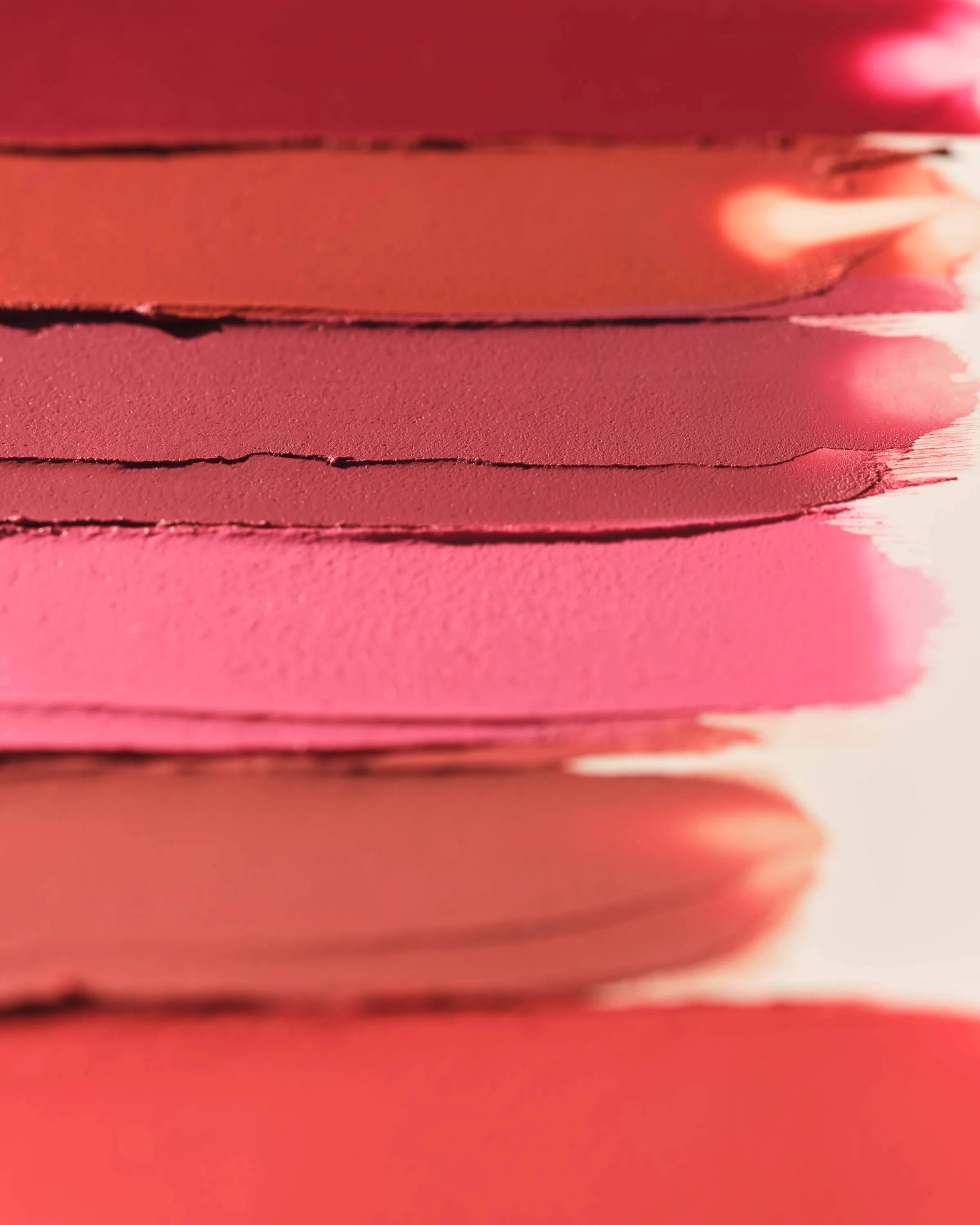 Close-up of red, coral and pink lipstick swatches smeared in stacked stripes on a cream surface, for Kylie Cosmetics.