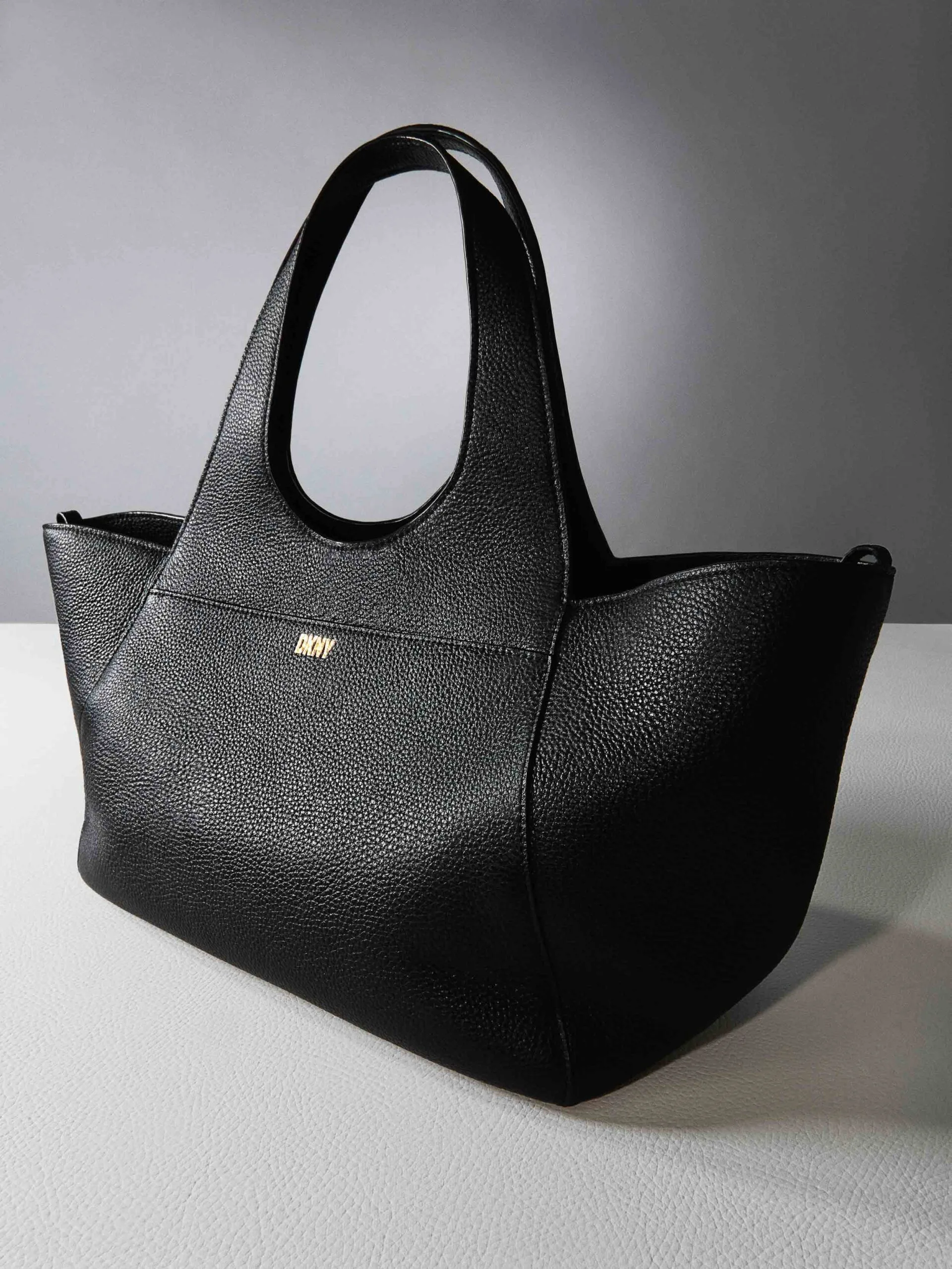 Black pebbled leather tote with gold logo and looped handles on a white surface against a grey backdrop, for DKNY.