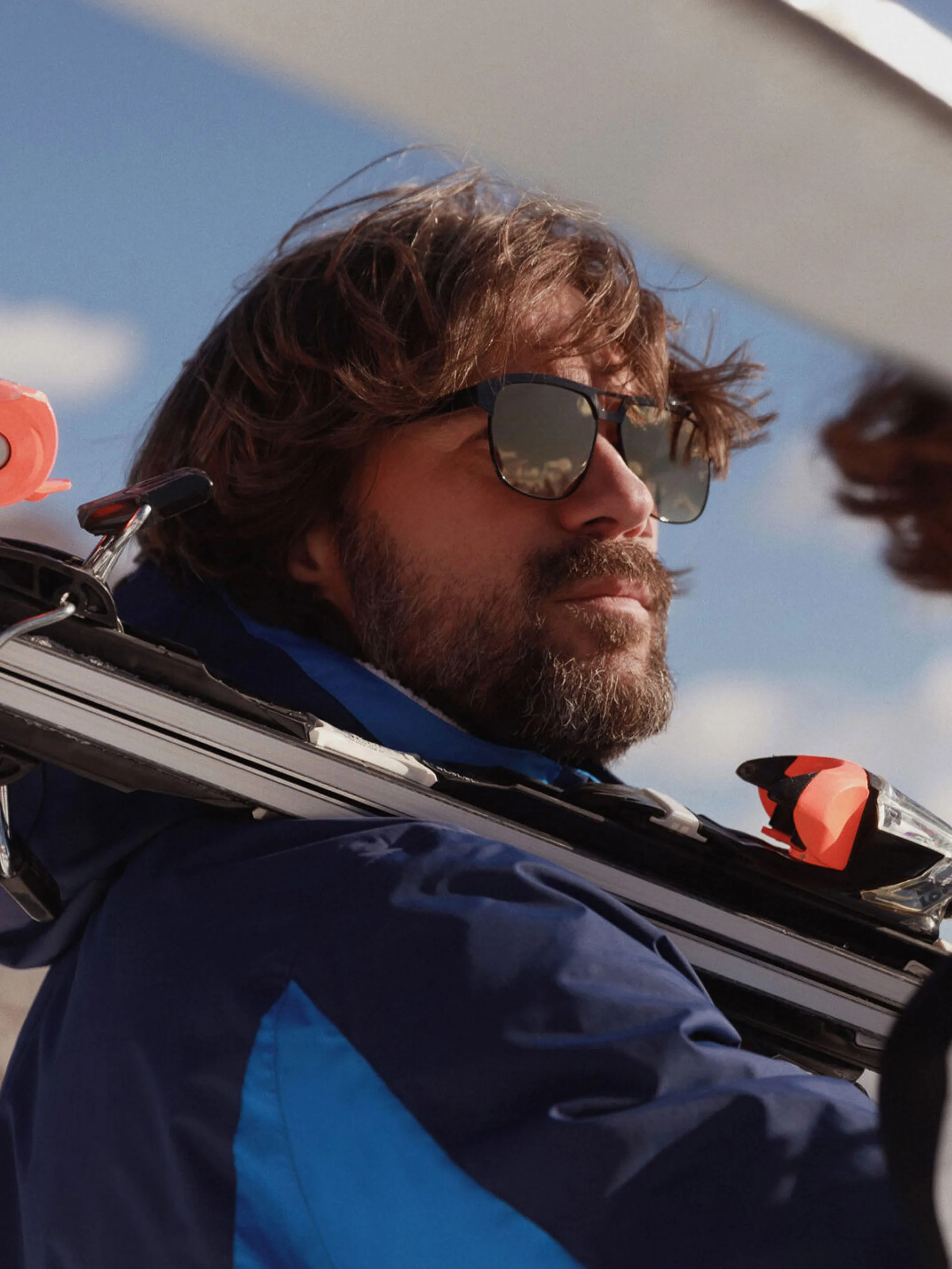Bearded man in mirrored sunglasses and a blue ski jacket carrying skis on his shoulder under a blue sky, for Marriott.