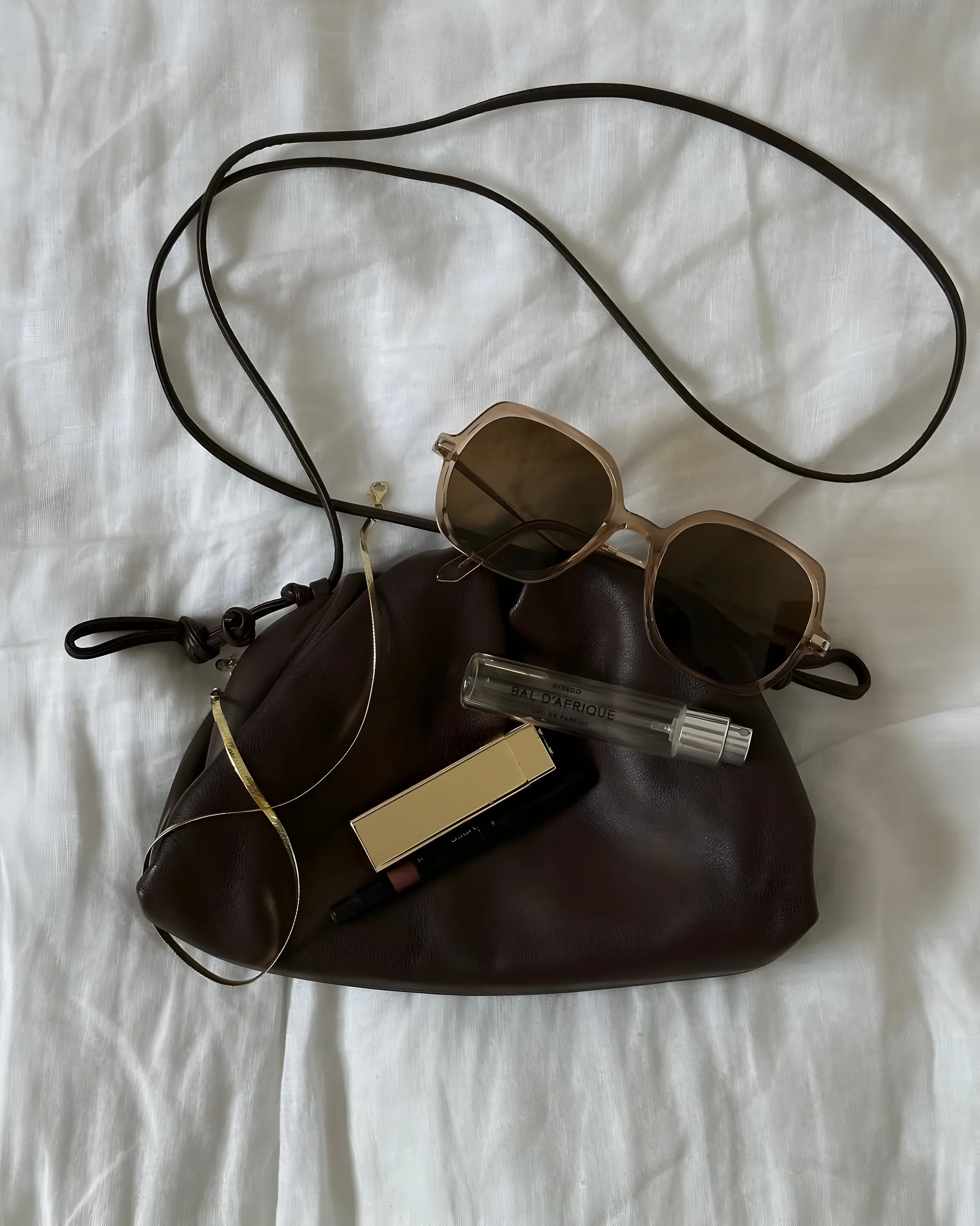 Brown leather pouch bag with sunglasses, Byredo Bal d'Afrique perfume, gold lipstick and necklace on white linen.