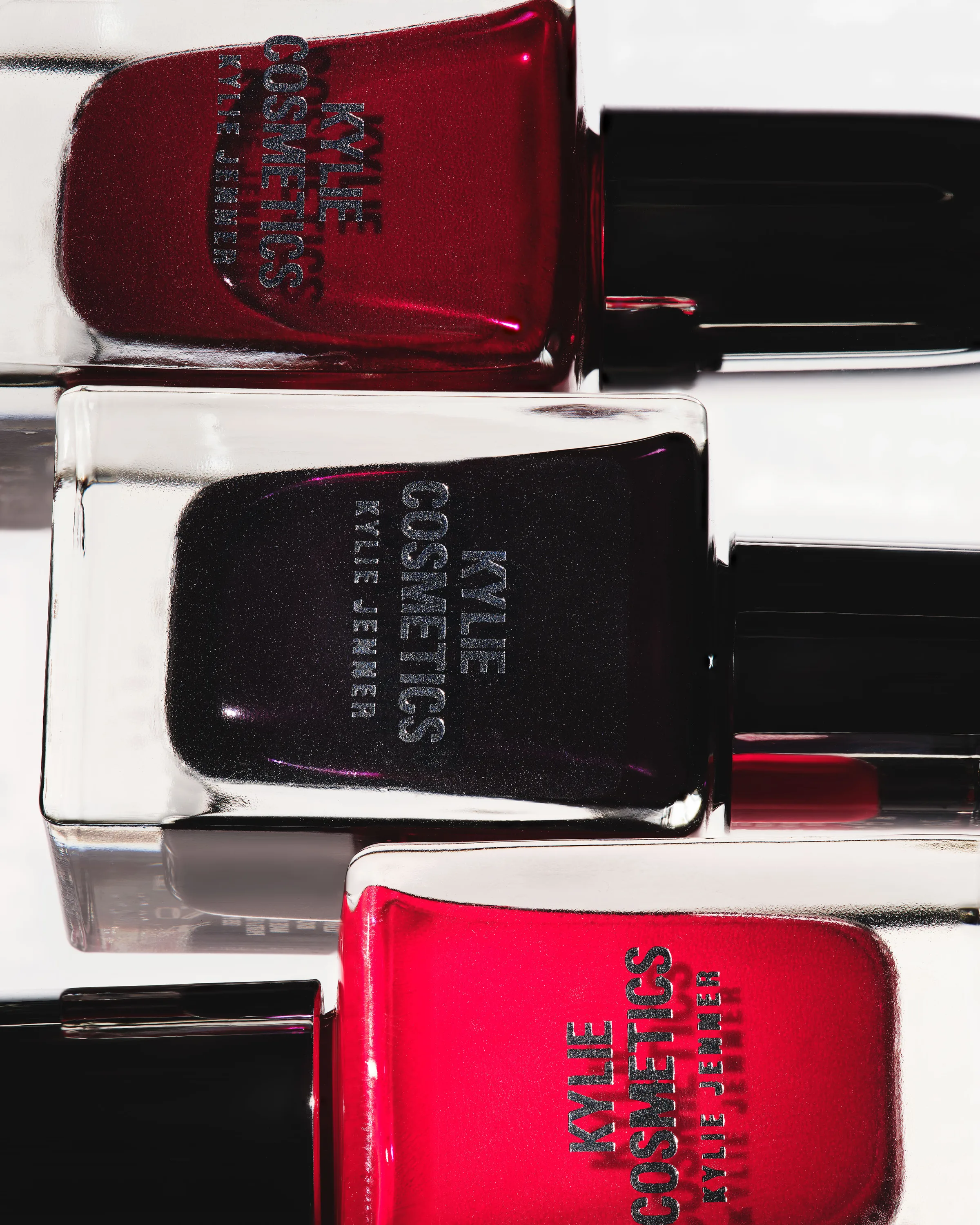 Three nail lacquer bottles in deep red, black and bright red lying stacked on a white surface, for Kylie Cosmetics.