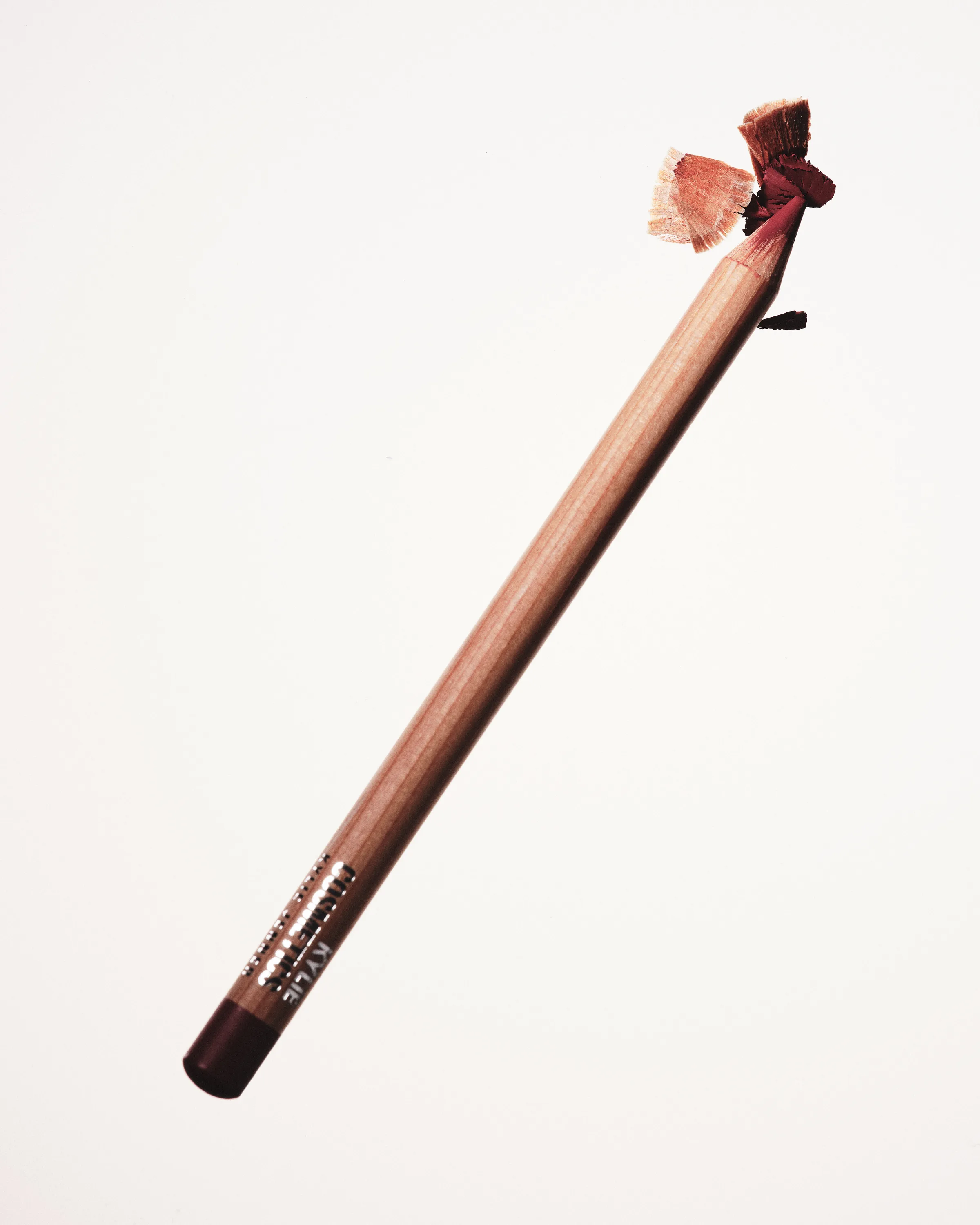 Wooden lip liner pencil in mauve with fresh shavings curling from its sharpened tip on white, for Kylie Cosmetics.