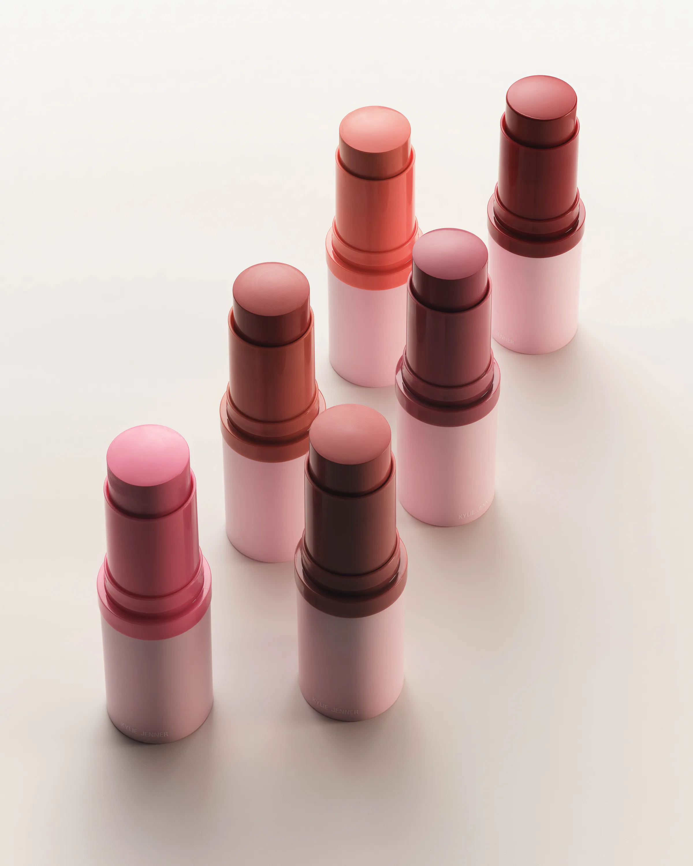 Six cream blush sticks in pink to berry shades with pink tubes, standing in diagonal rows on white, for Kylie Cosmetics.