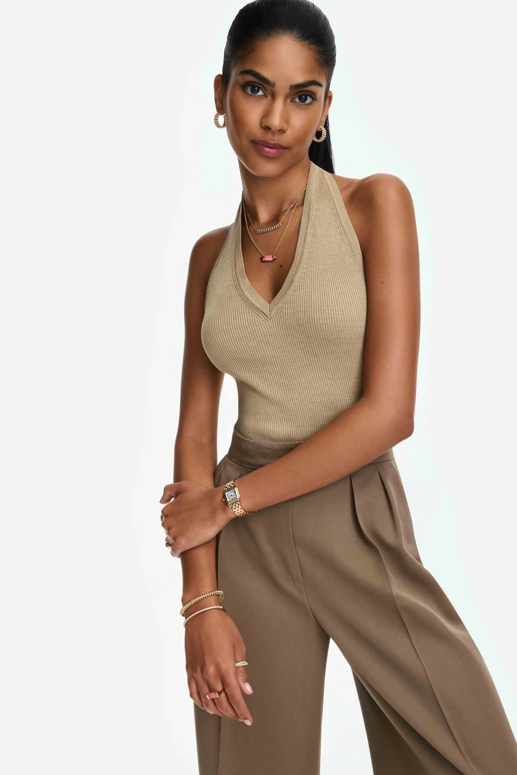 Model in beige ribbed halter top and taupe trousers wearing stacked gold bracelets, watch and necklaces, for Cartier.