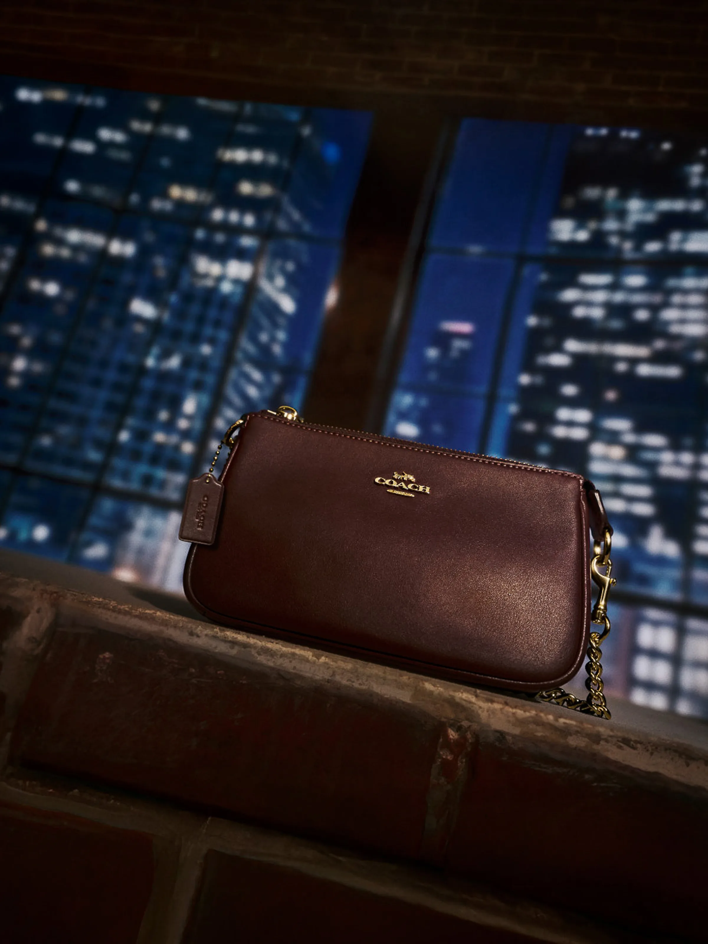 AI image of a burgundy leather wristlet with gold chain on a brick ledge before night city windows, for Coach.
