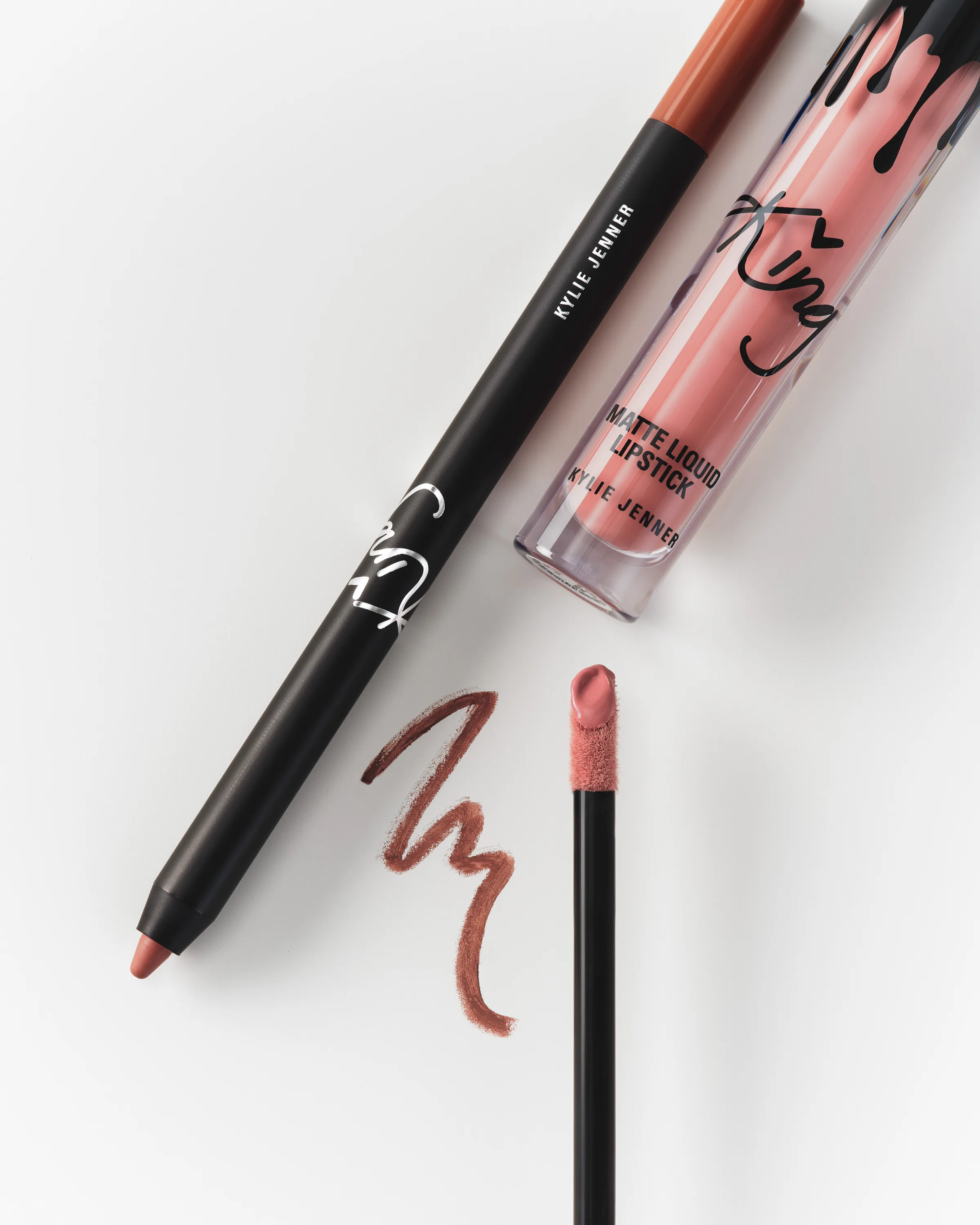 Black lip liner and King matte liquid lipstick with applicator and brown swatch on white, still life, for Kylie Cosmetics.