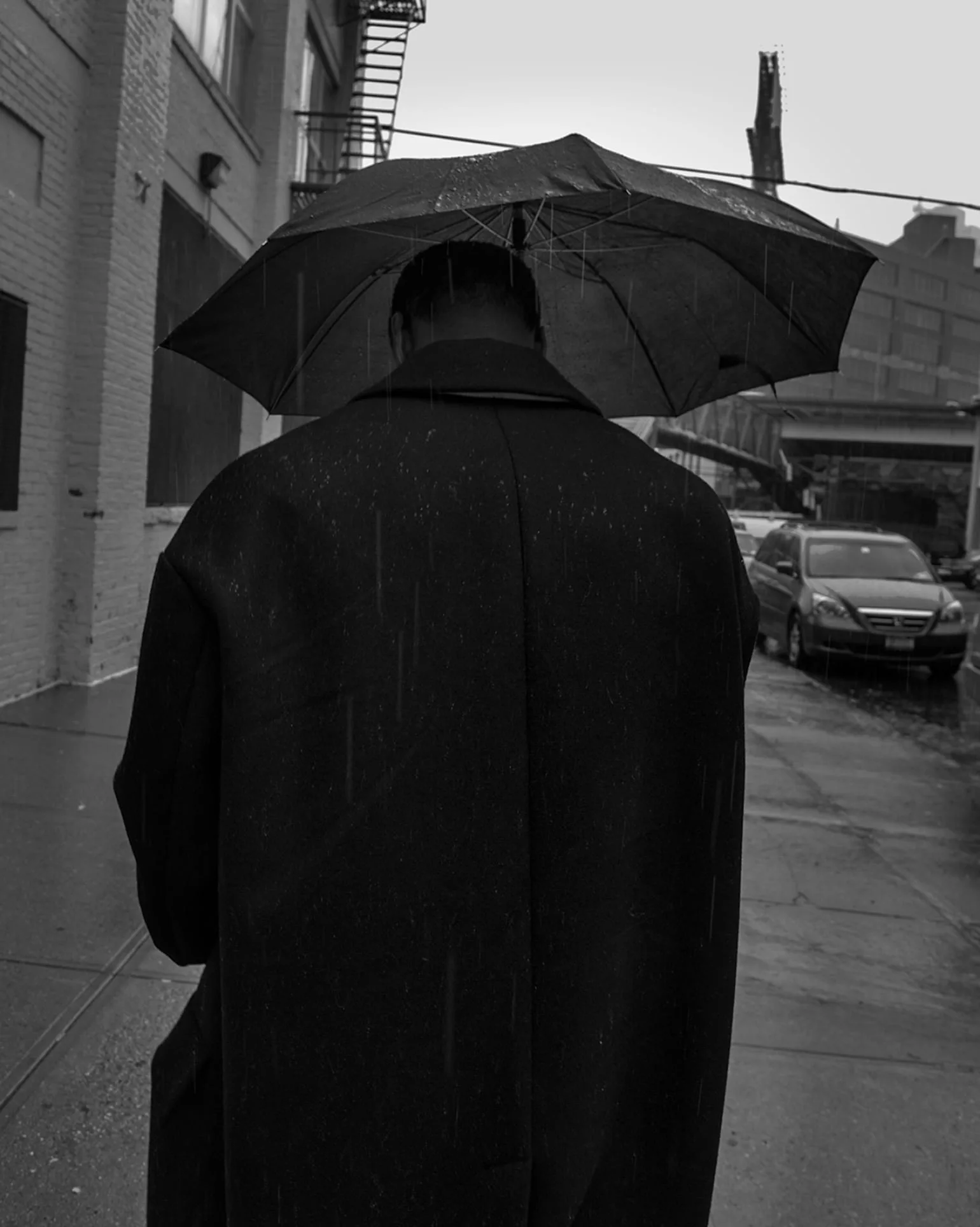 Black and white shot of a man in a dark overcoat walking under an umbrella on a rainy street, for The Last Magazine.
