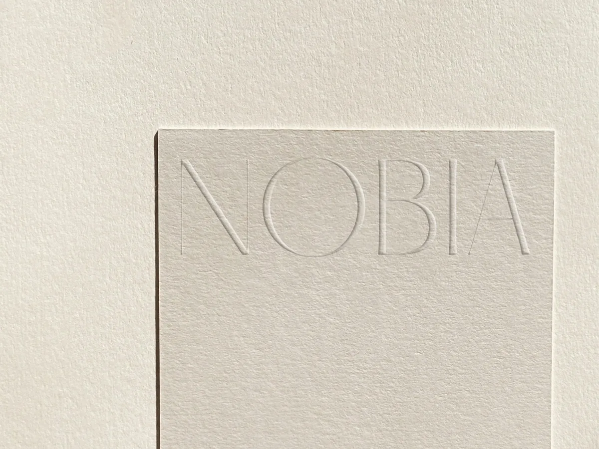 Cream textured paper card with the NOBIA wordmark blind-debossed across the top, on matching paper, for Nobia.