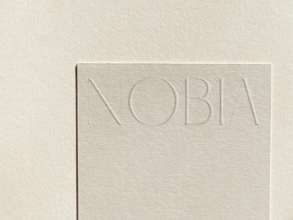 Cream textured paper card with the NOBIA wordmark blind-debossed across the top, on matching paper, for Nobia.