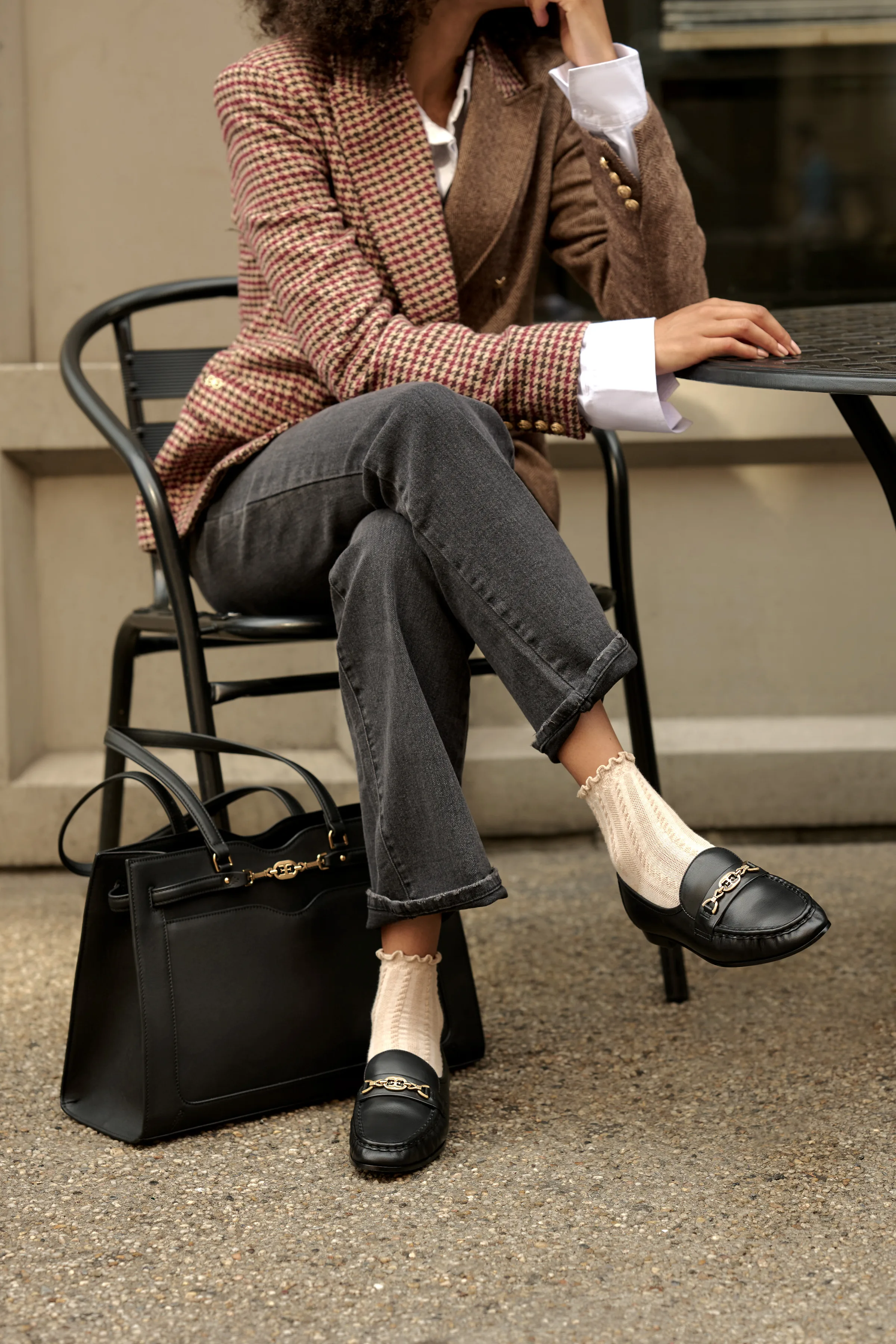 Woman in houndstooth blazer and grey jeans seated at café table with black chain-trim loafers and tote, for Sam Edelman.