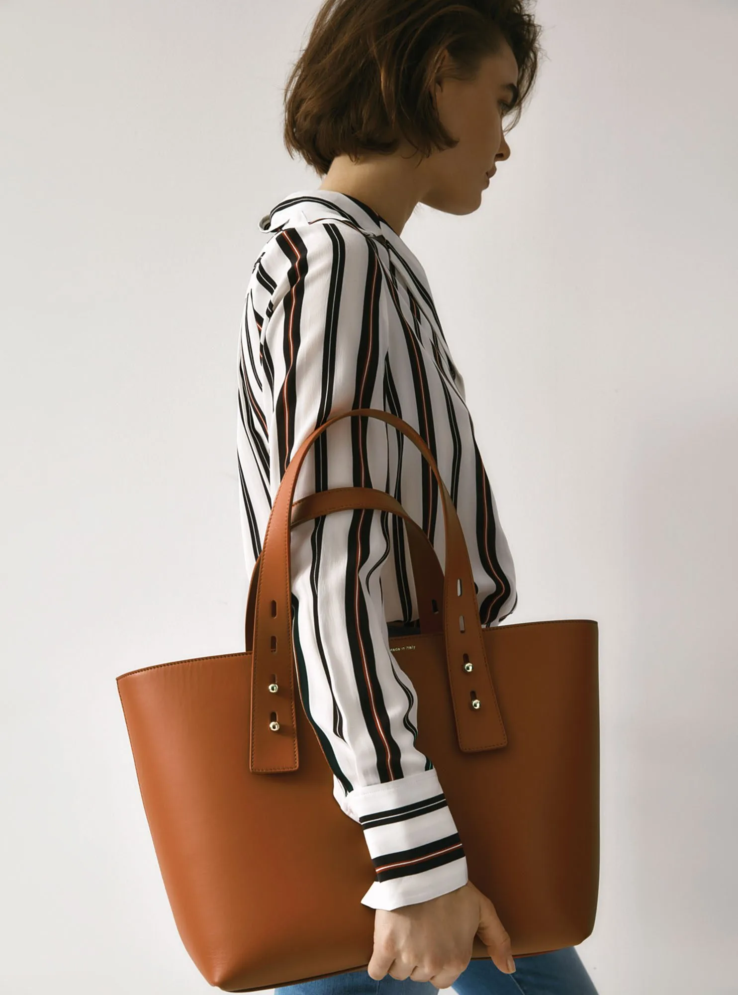 Model in a black and white striped shirt carrying a cognac leather tote bag against a white wall, for FRAME.