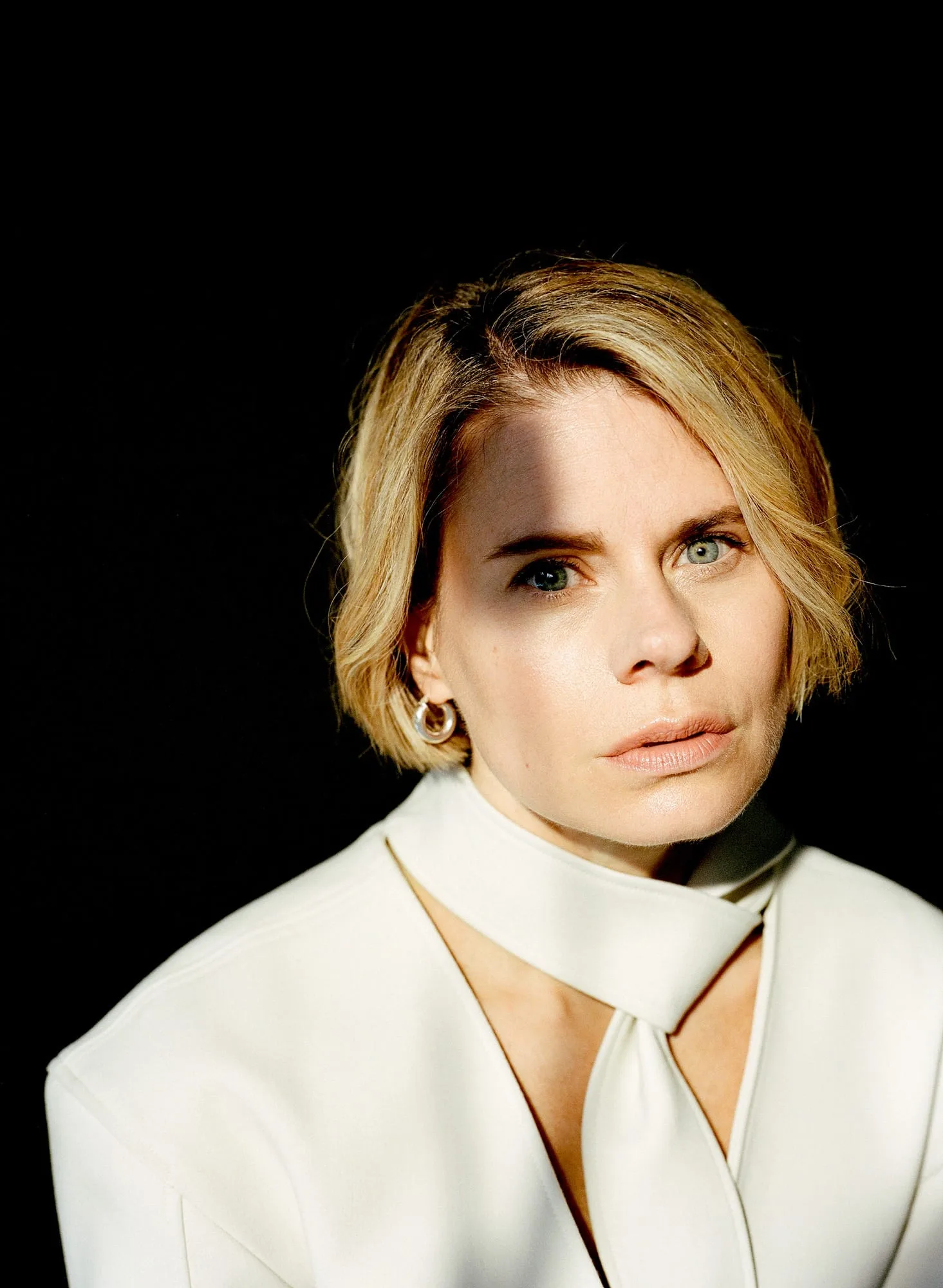 Portrait of a woman with a blonde bob in a white tie-neck top, lit by a slash of sunlight on black, for The Last Magazine.