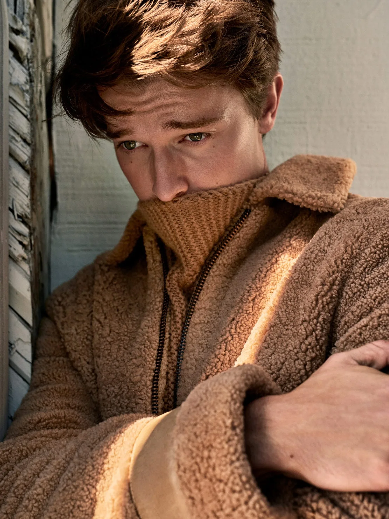 Young man in a camel shearling jacket and ribbed turtleneck leaning against a weathered wall, for Man About Town.